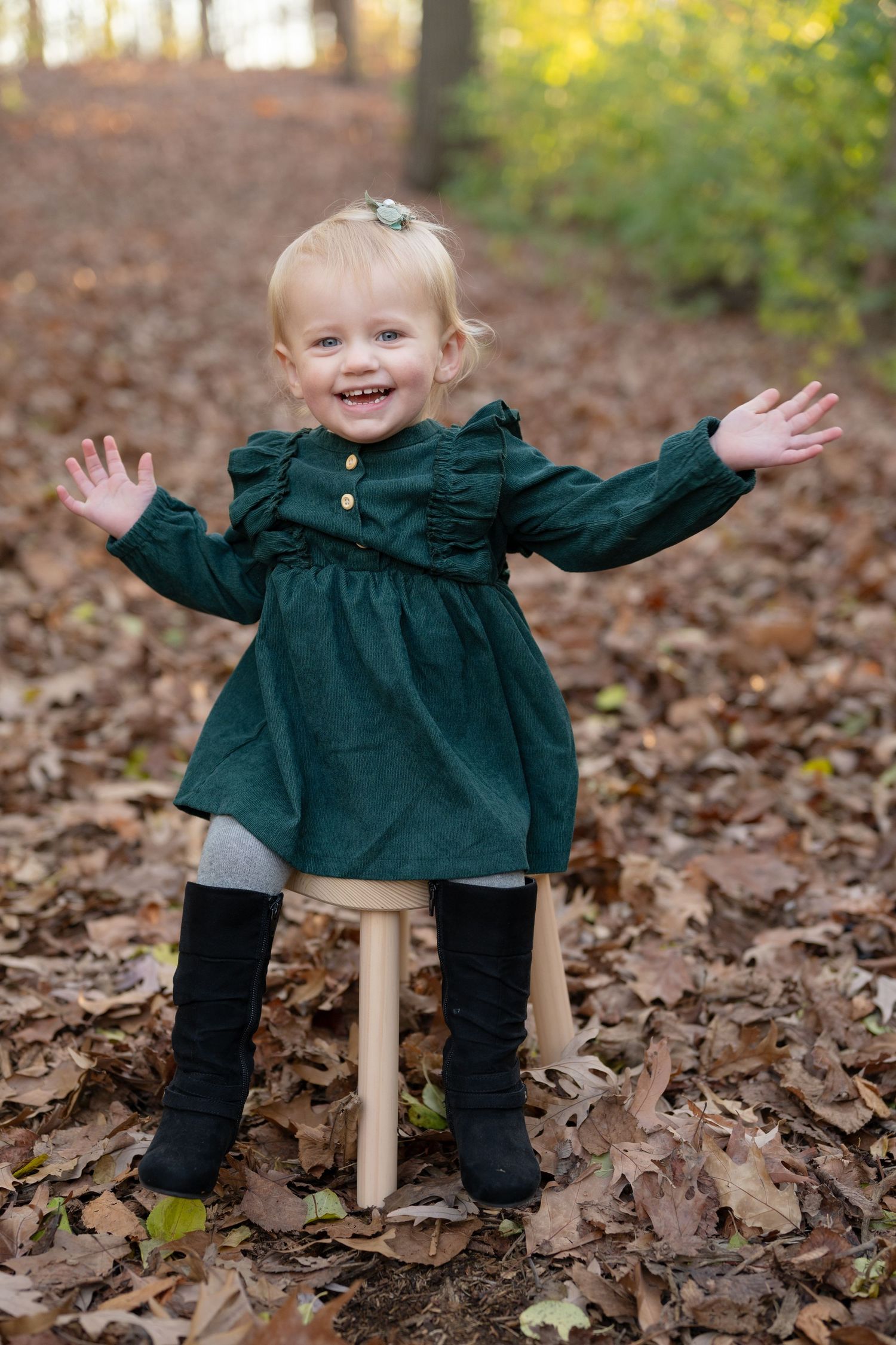 Toddler in green dress with arms outstretched standing on wooden stool among fall leaves.