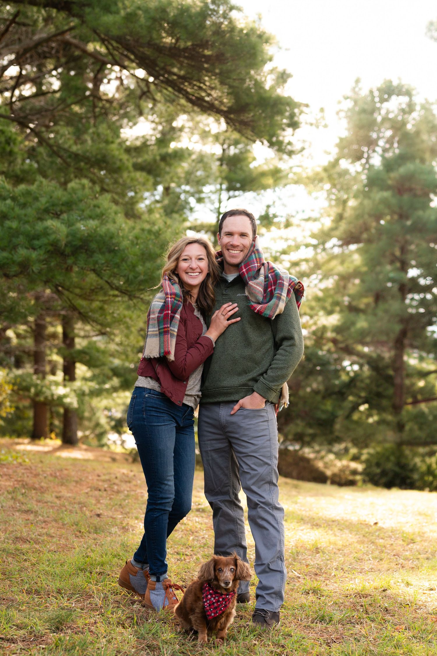 Couple in casual fall attire poses with small dog in sunlit forest setting.