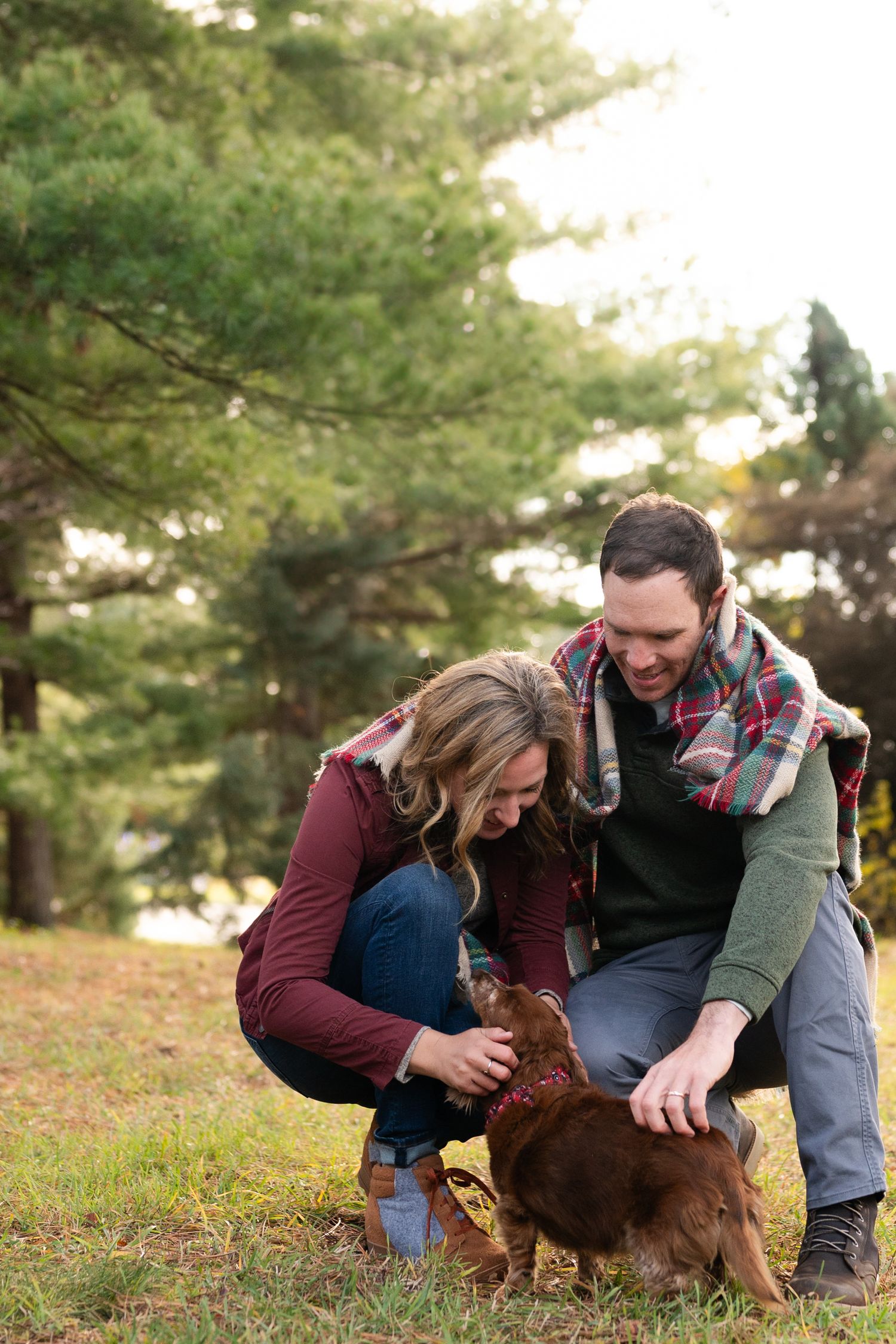 A couple kneels on autumn ground petting a brown dog in a wooded setting.