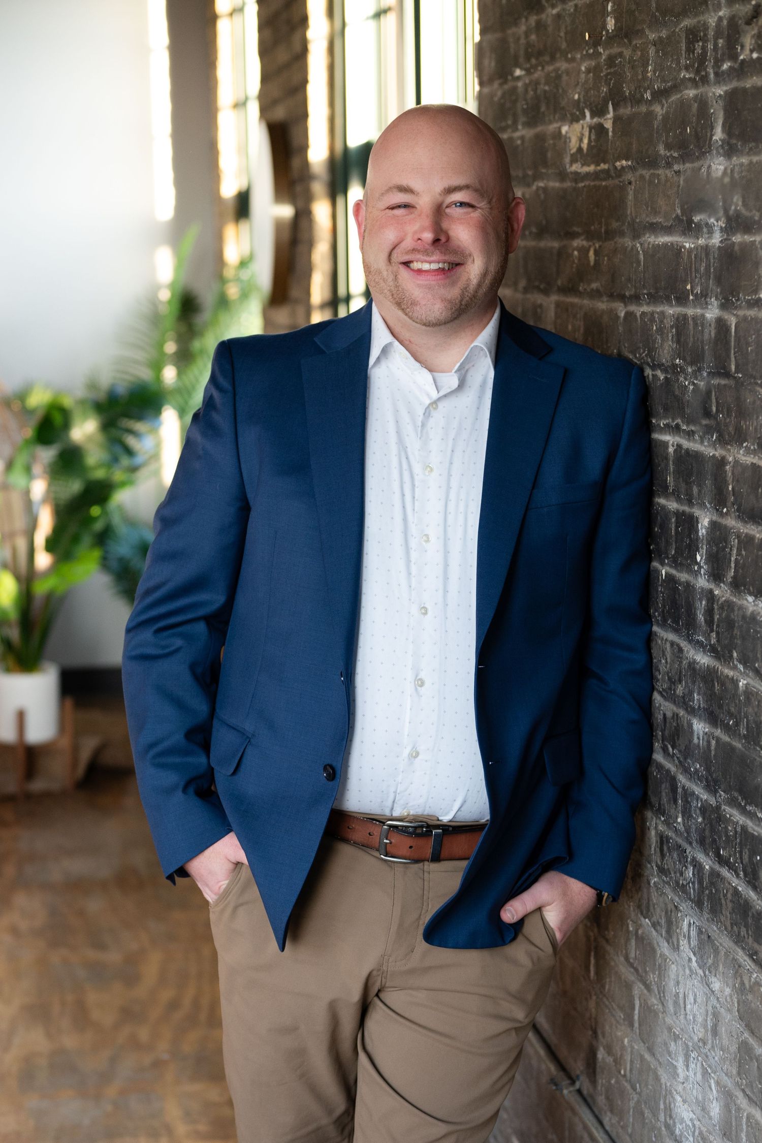 Professional in navy suit jacket poses against brick wall in modern industrial space with natural lighting.