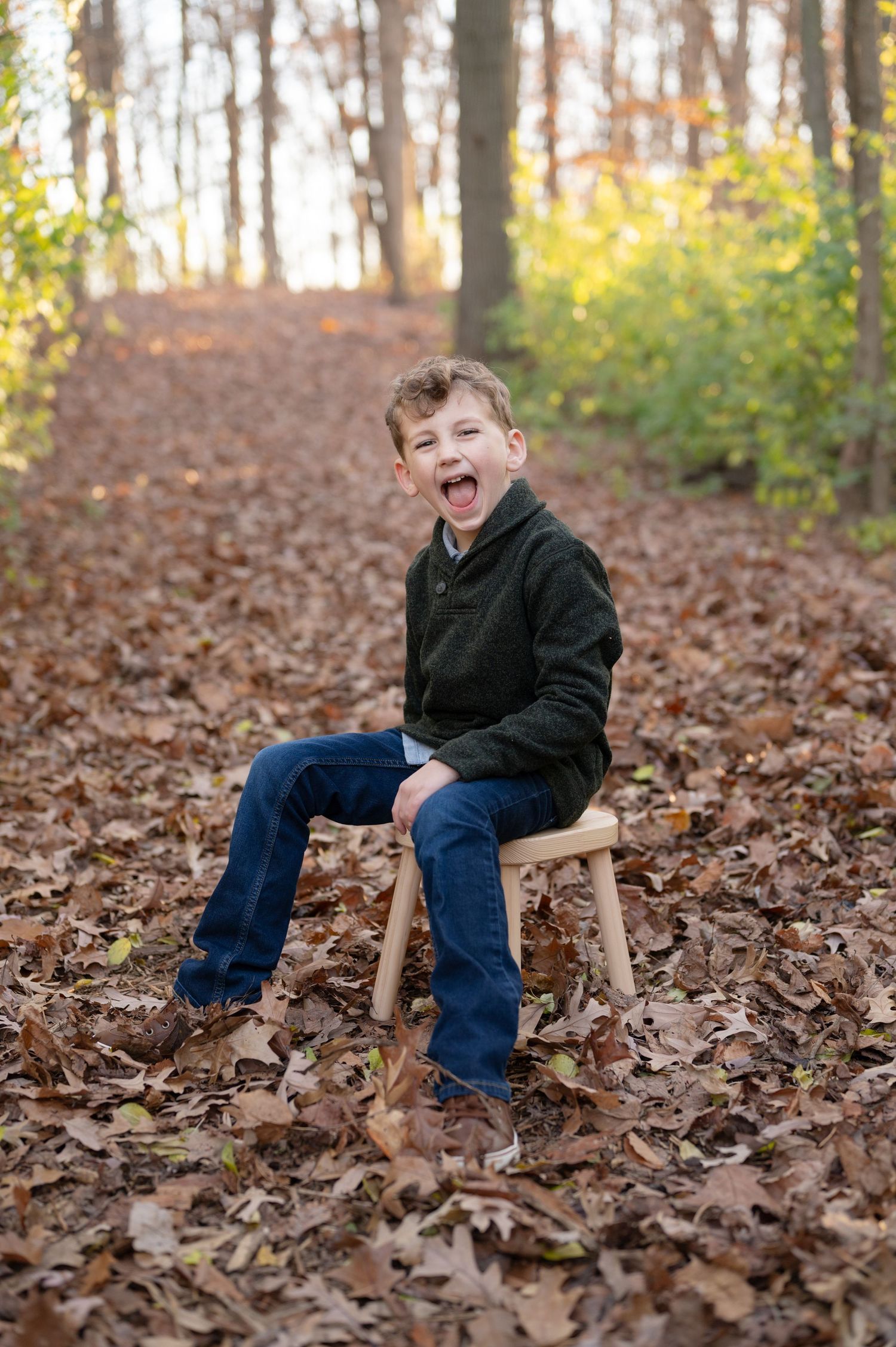 Child sitting on wooden stool surrounded by autumn leaves in the forest.