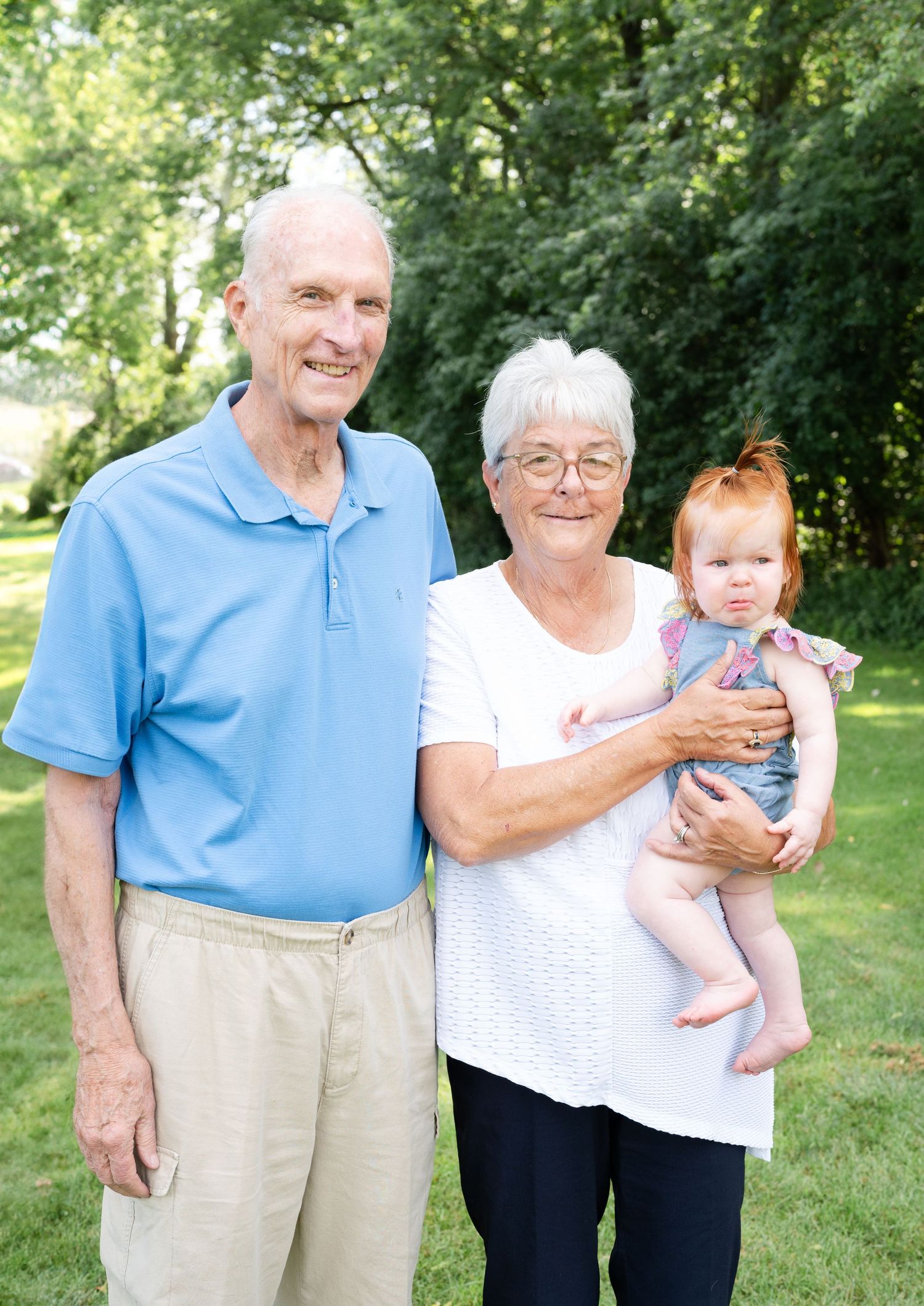 Three generations pose together outdoors with a small baby on a sunny summer day.