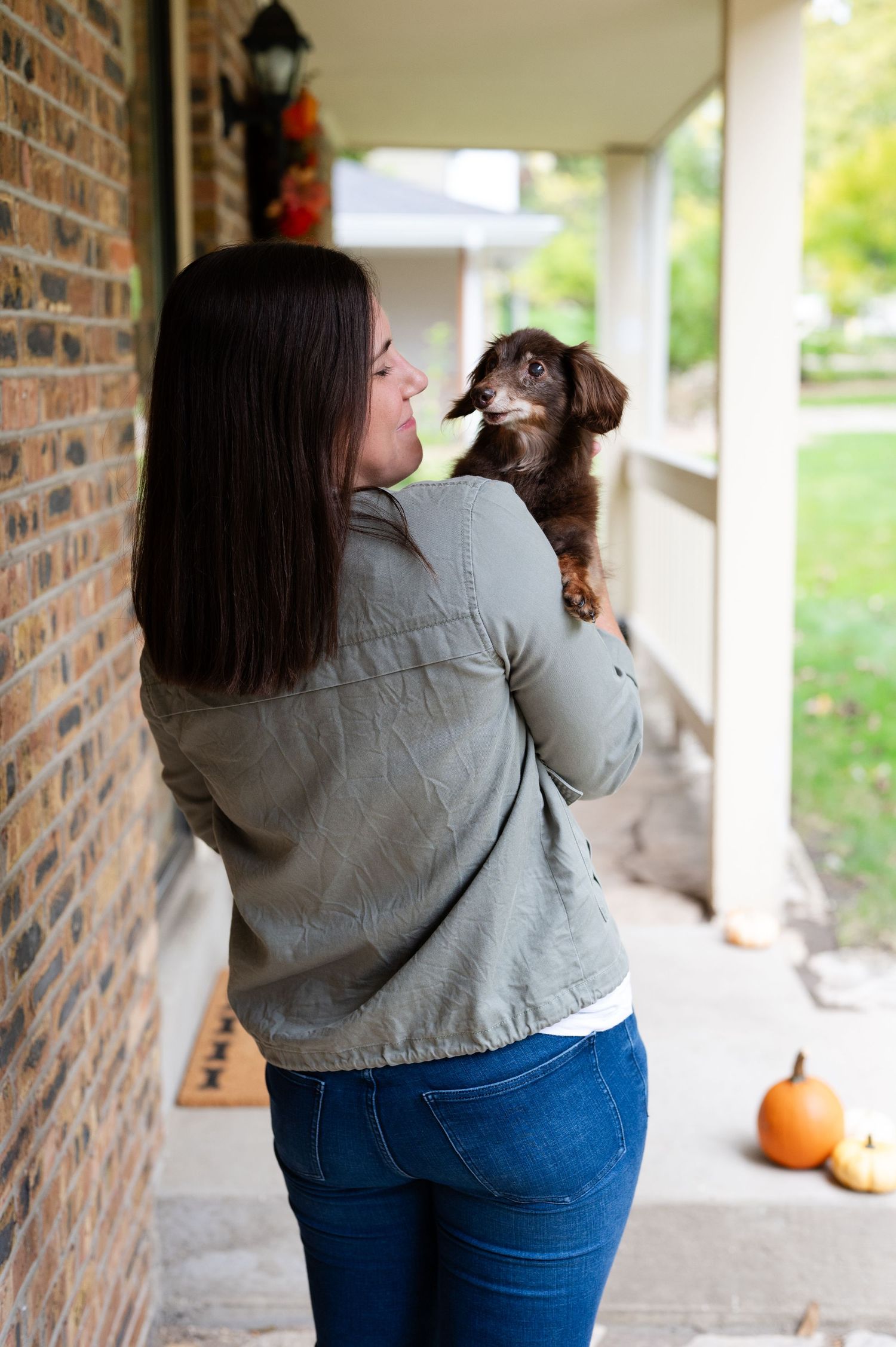 A brown dachshund puppy being held on a porch with fall decorations visible.