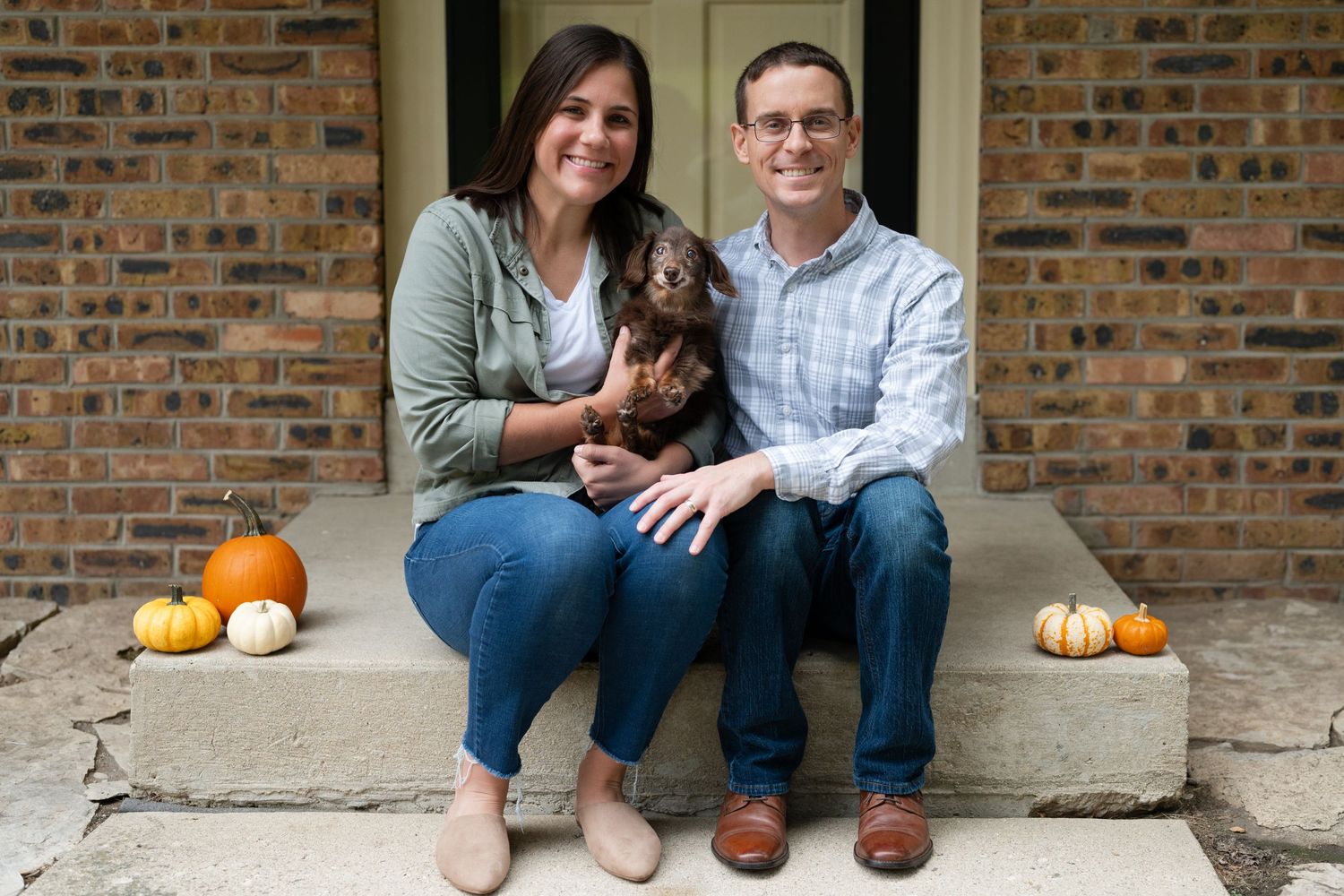 Couple sitting on front porch steps with dachshund dog and autumn pumpkin decorations.