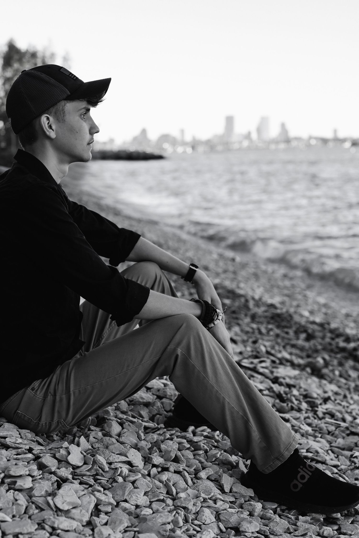 Black and white portrait of person in baseball cap sitting on rocky shoreline with cityscape in background.
