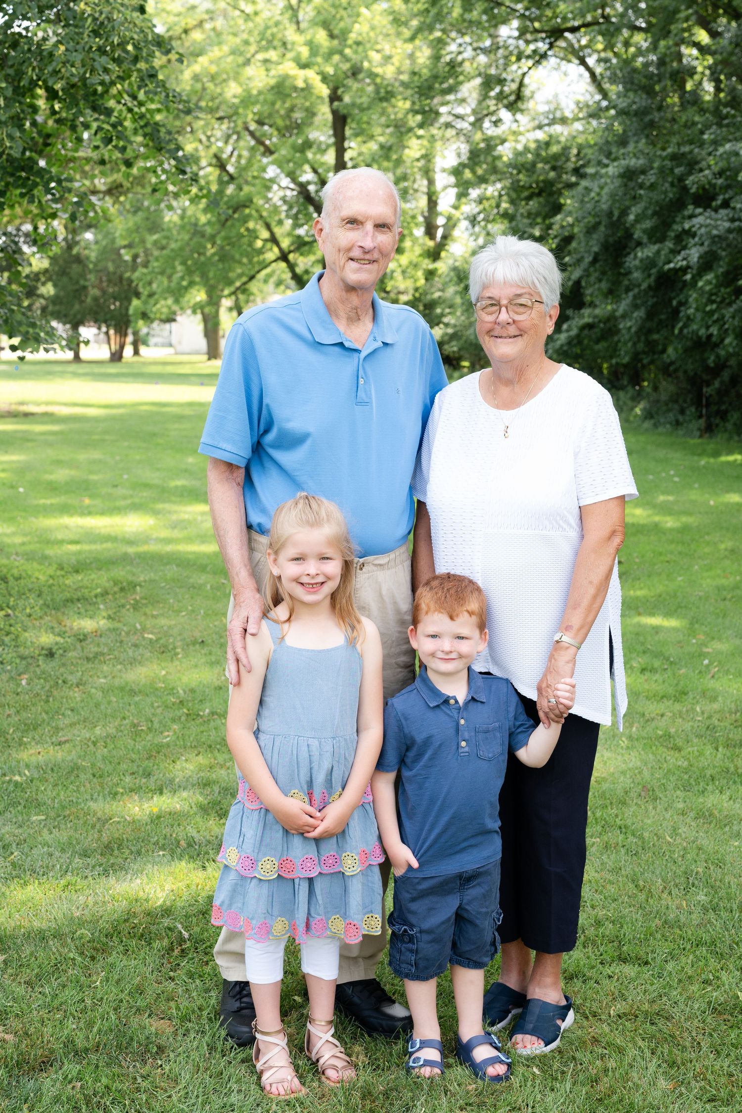 A family group of four poses outdoors on a sunny day in a park with green trees in the background.