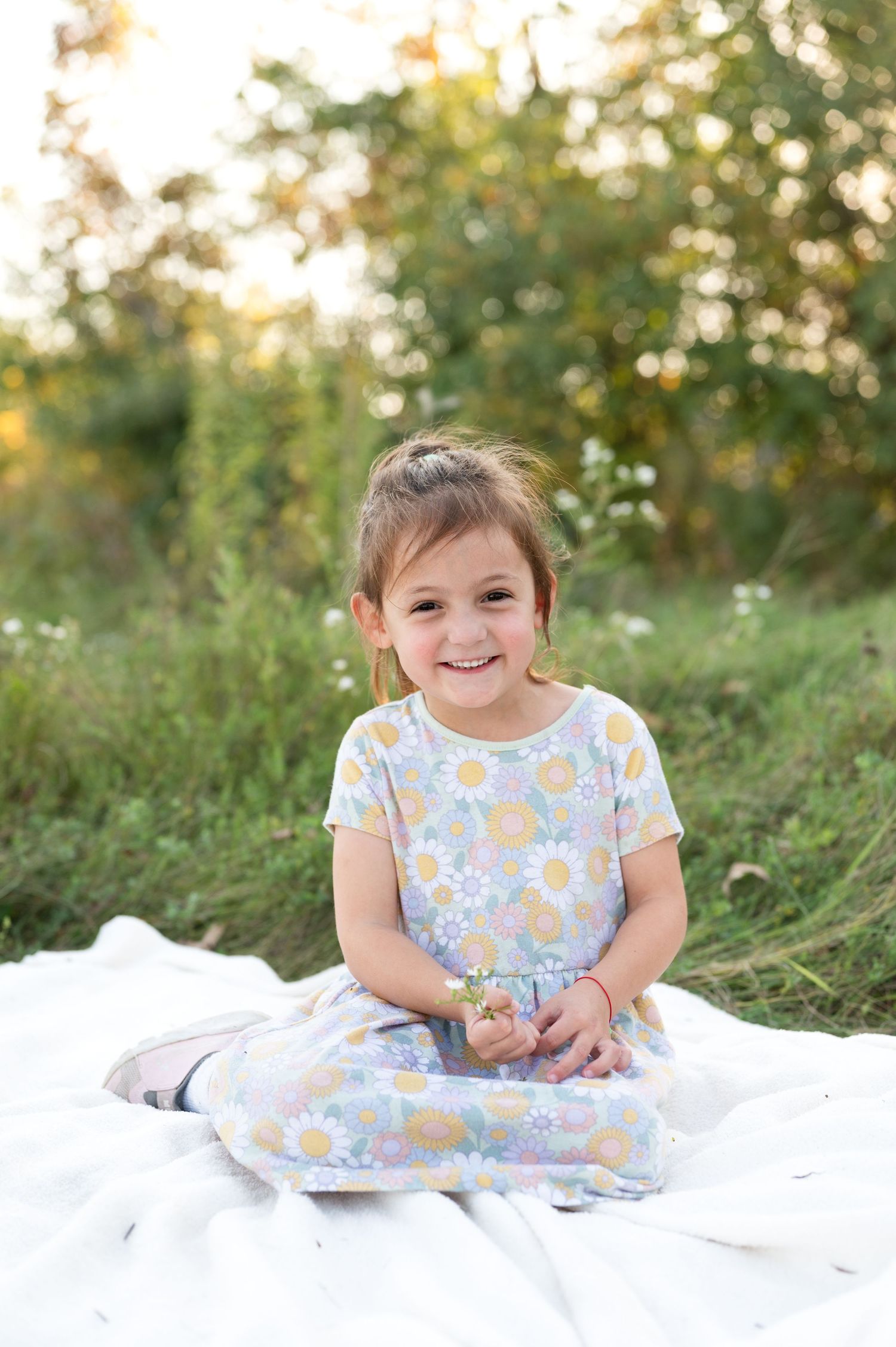 A child in a floral dress sits on a white blanket outdoors during a sunny summer photoshoot.