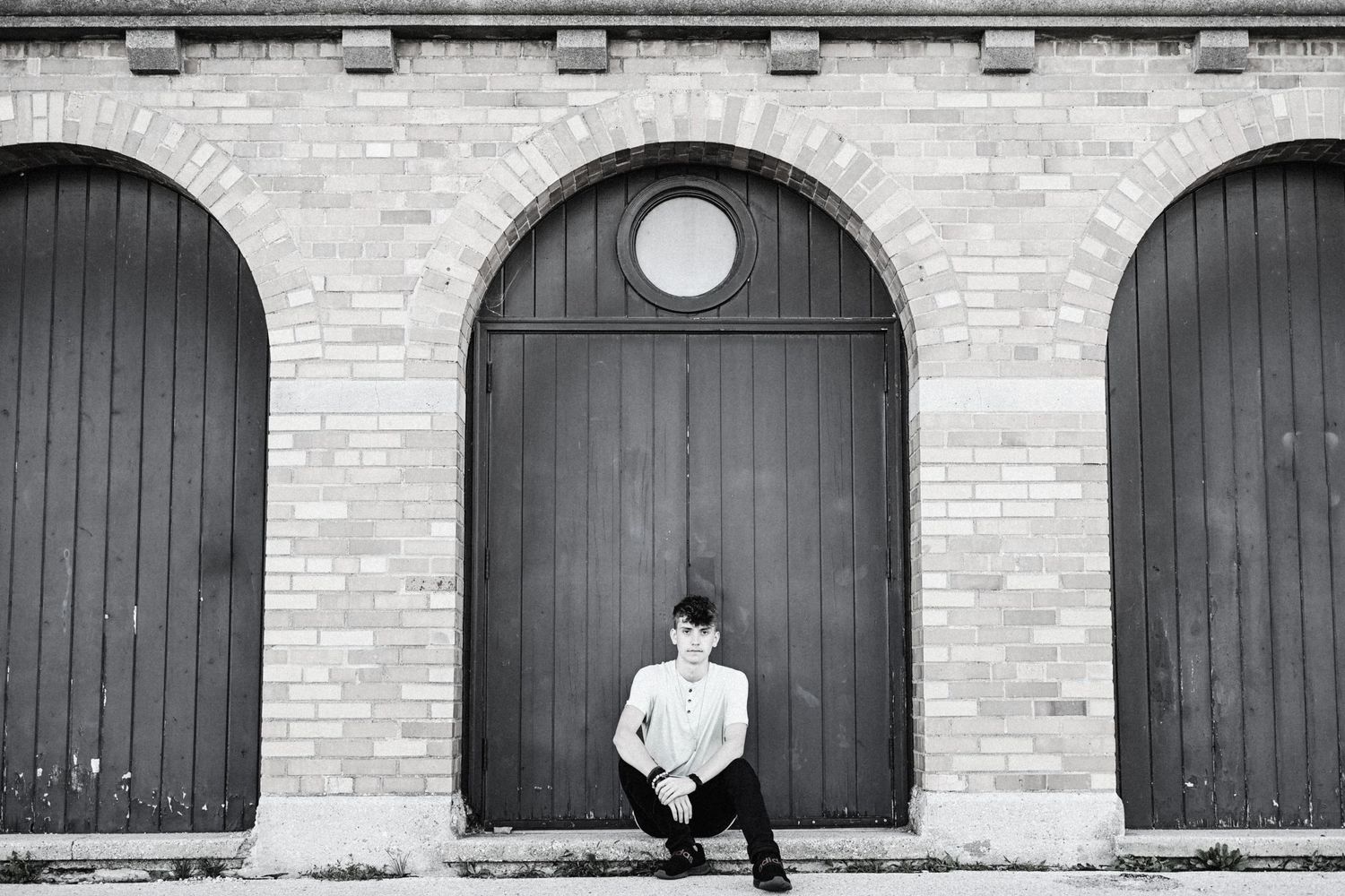Black and white photo of someone seated in front of three arched doorways with brick walls.