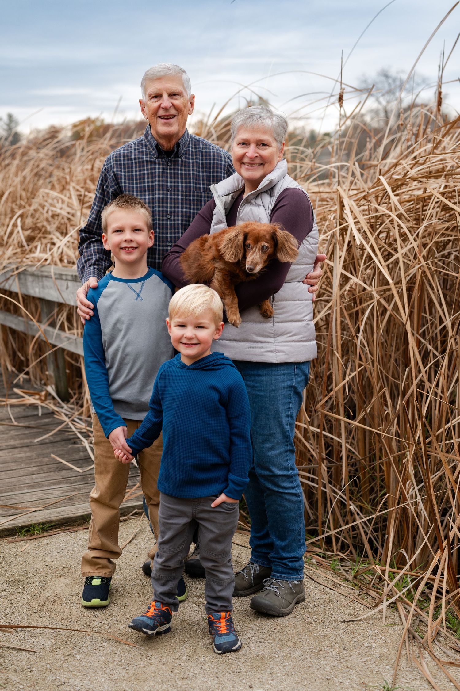 Family portrait on wooden boardwalk with their brown dog among tall grass.