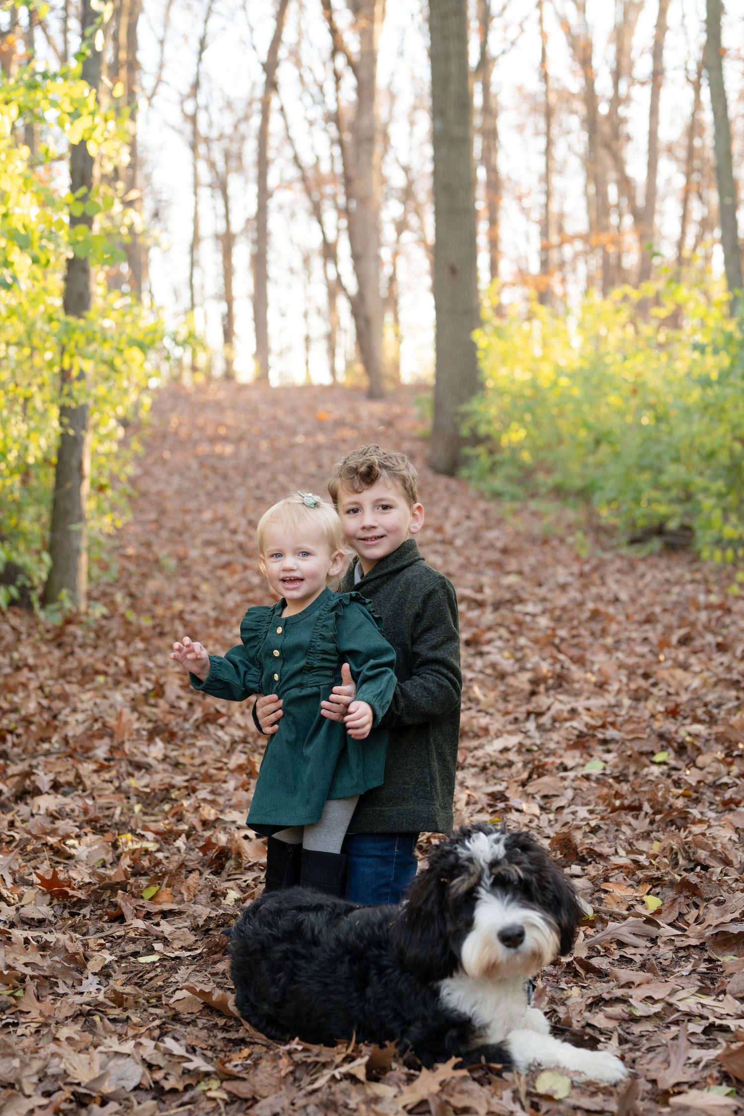 Two children standing with black and white dog on leaf-covered forest path.