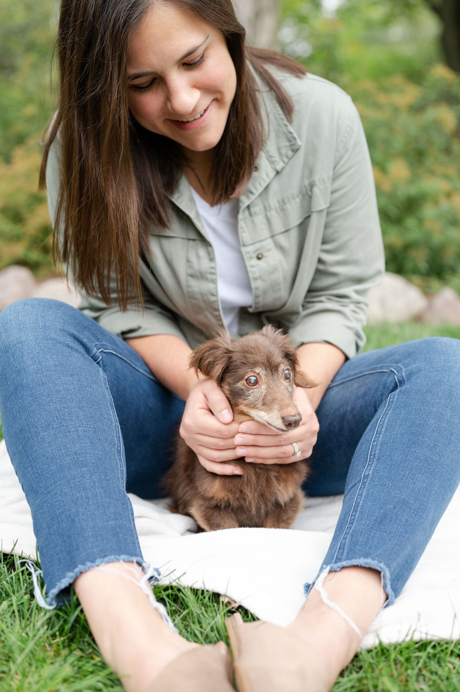 Woman in casual attire sitting outdoors on blanket petting small brown dachshund.