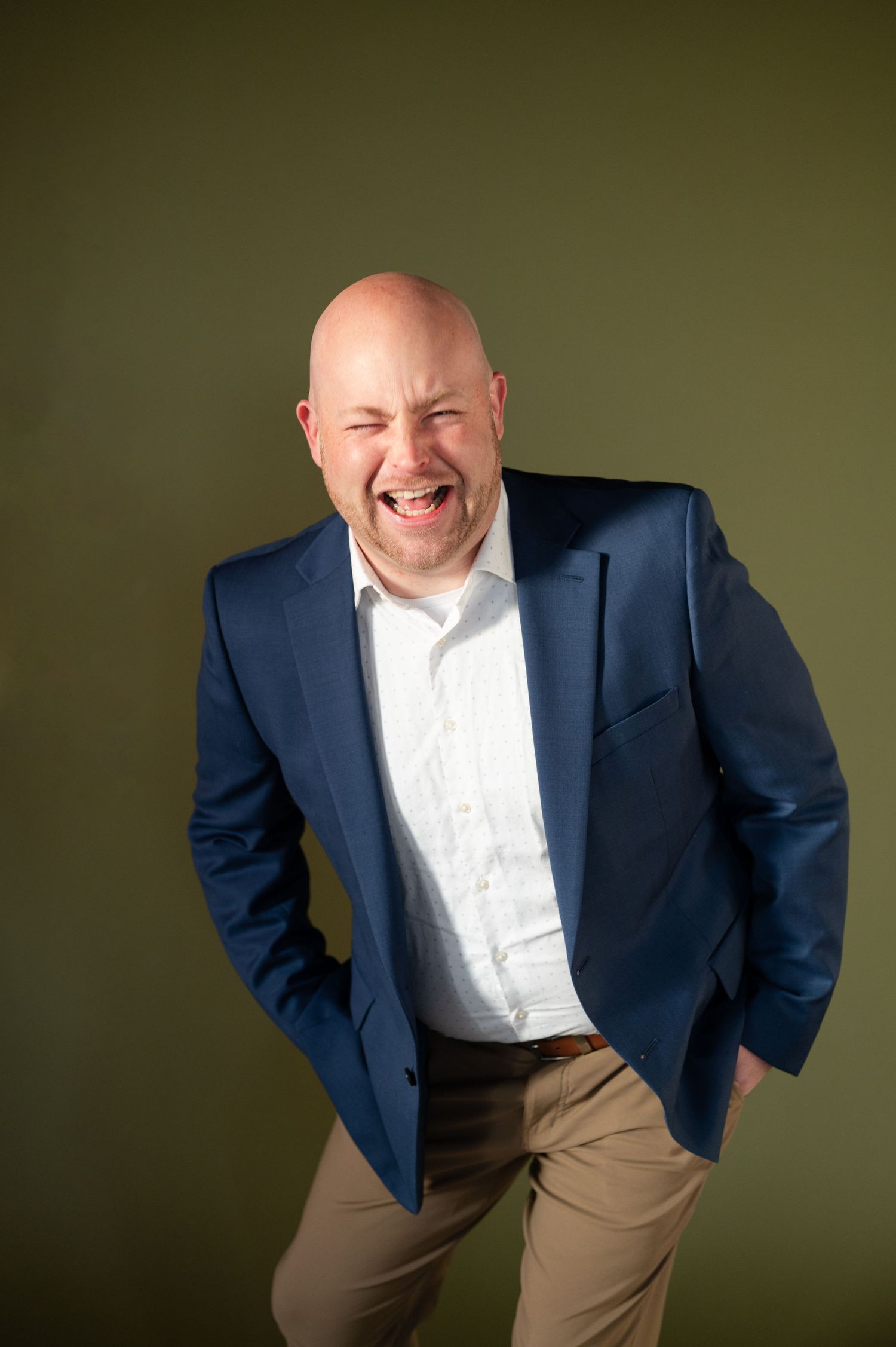 Professional in navy blazer and khakis poses casually against olive background with expressive smile.