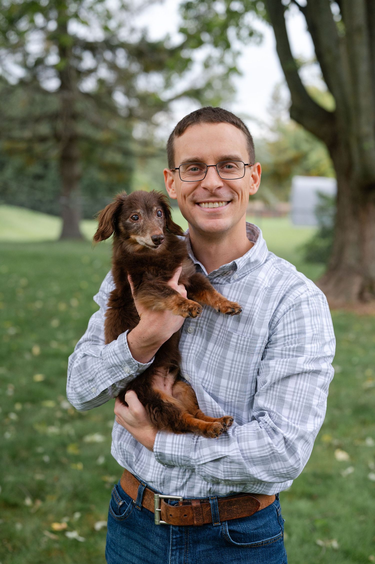 Man holding brown dachshund puppy while standing in park with trees in background.