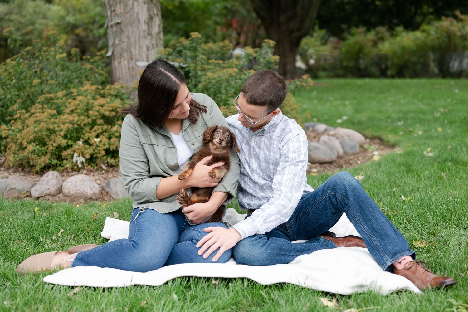 Couple sitting on blanket in park with small brown dog between them.