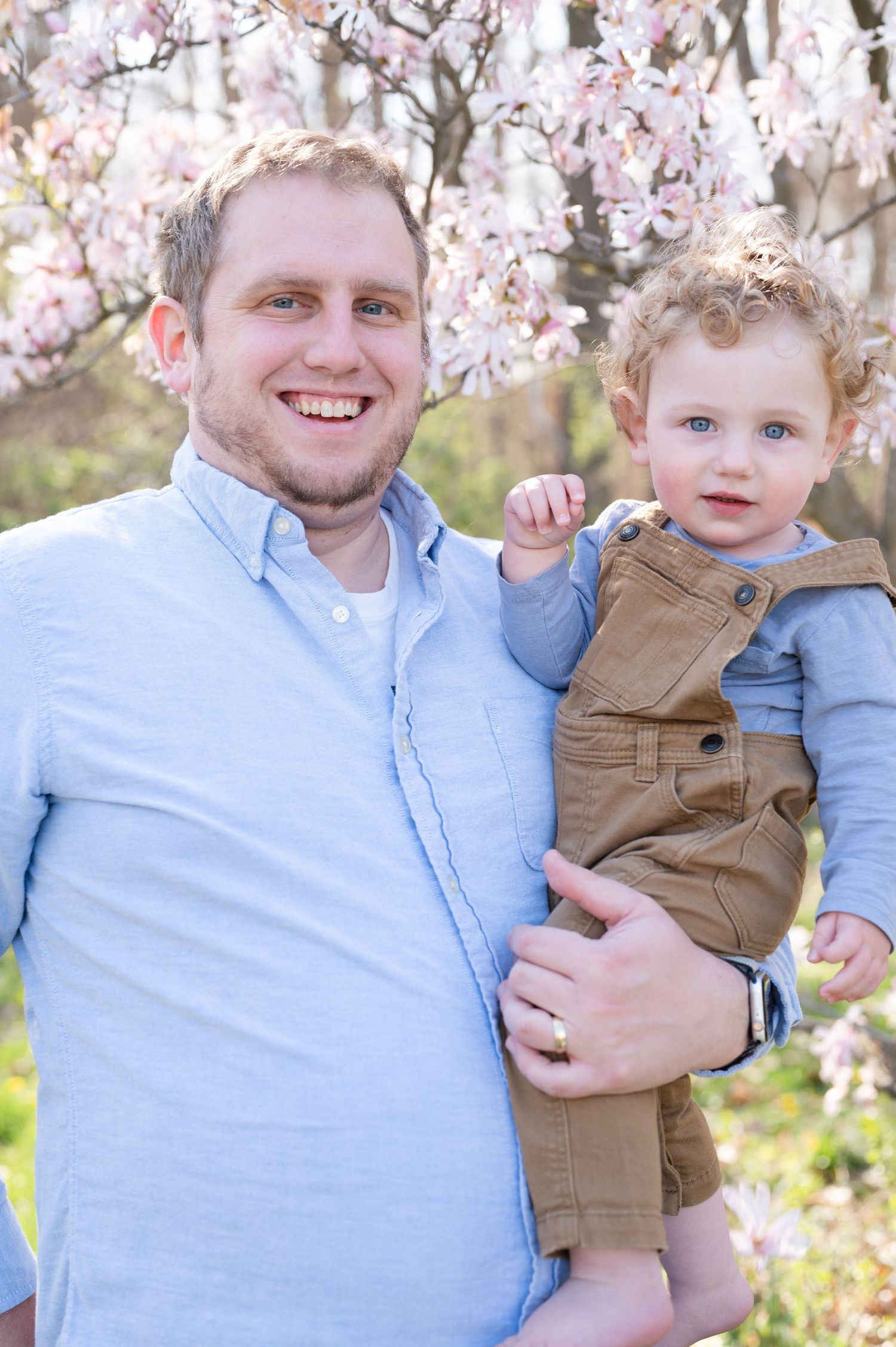 Joyful outdoor portrait with cherry blossoms featuring casual light blue shirt and brown overalls.