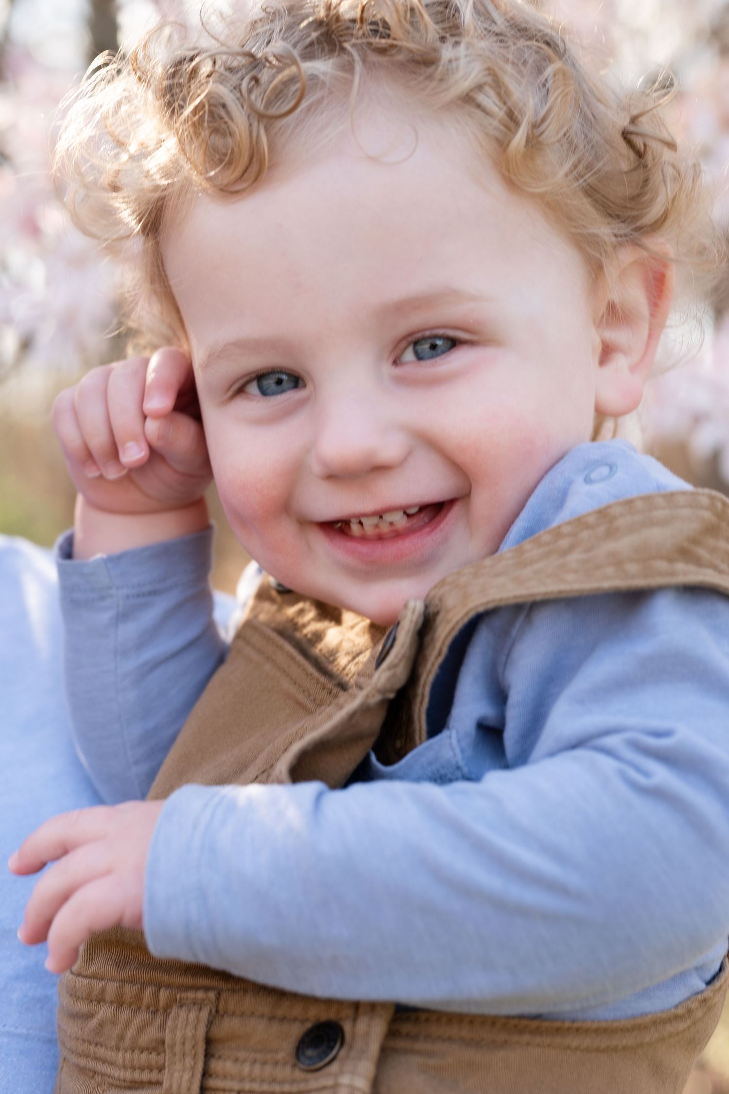 Young child in brown overalls smiling at camera with cherry blossoms in background.