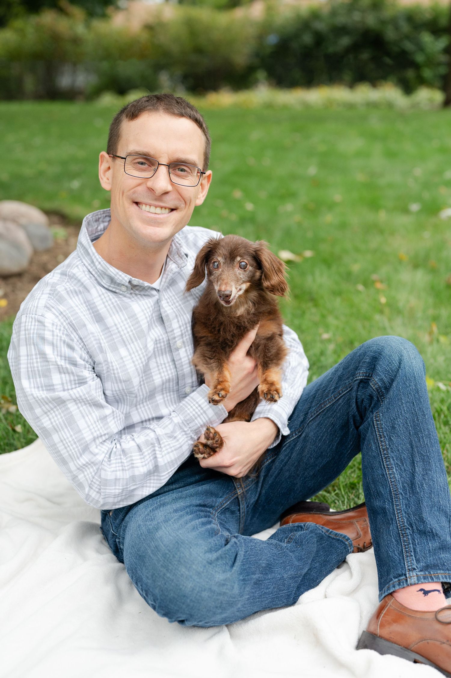 Person in checkered shirt holding small brown dachshund while sitting on white blanket outdoors.
