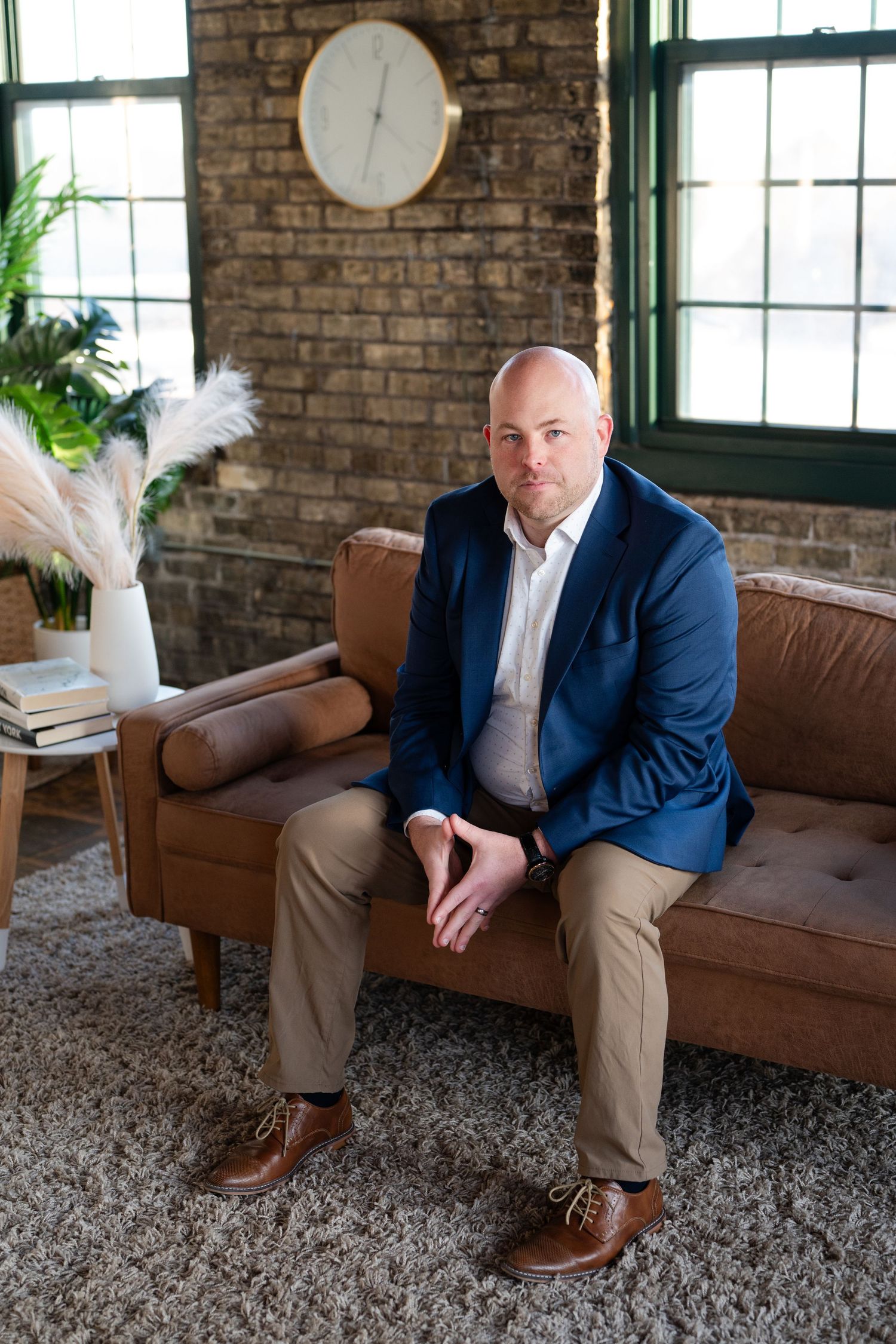Professional in business attire sits on brown leather couch in industrial-style room with exposed brick walls.