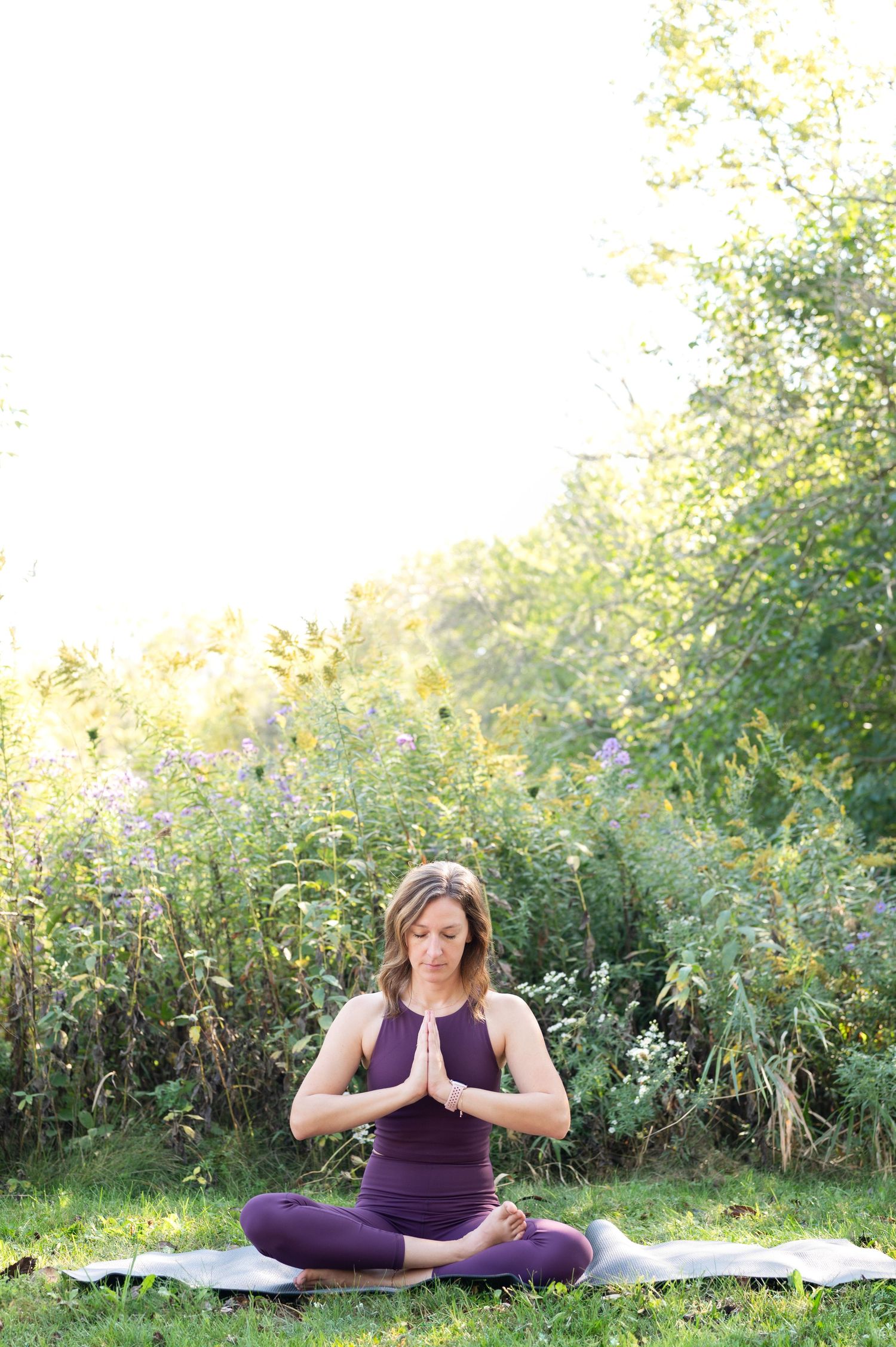 A person sits in lotus position on a yoga mat outdoors, practicing meditation among wildflowers and greenery.