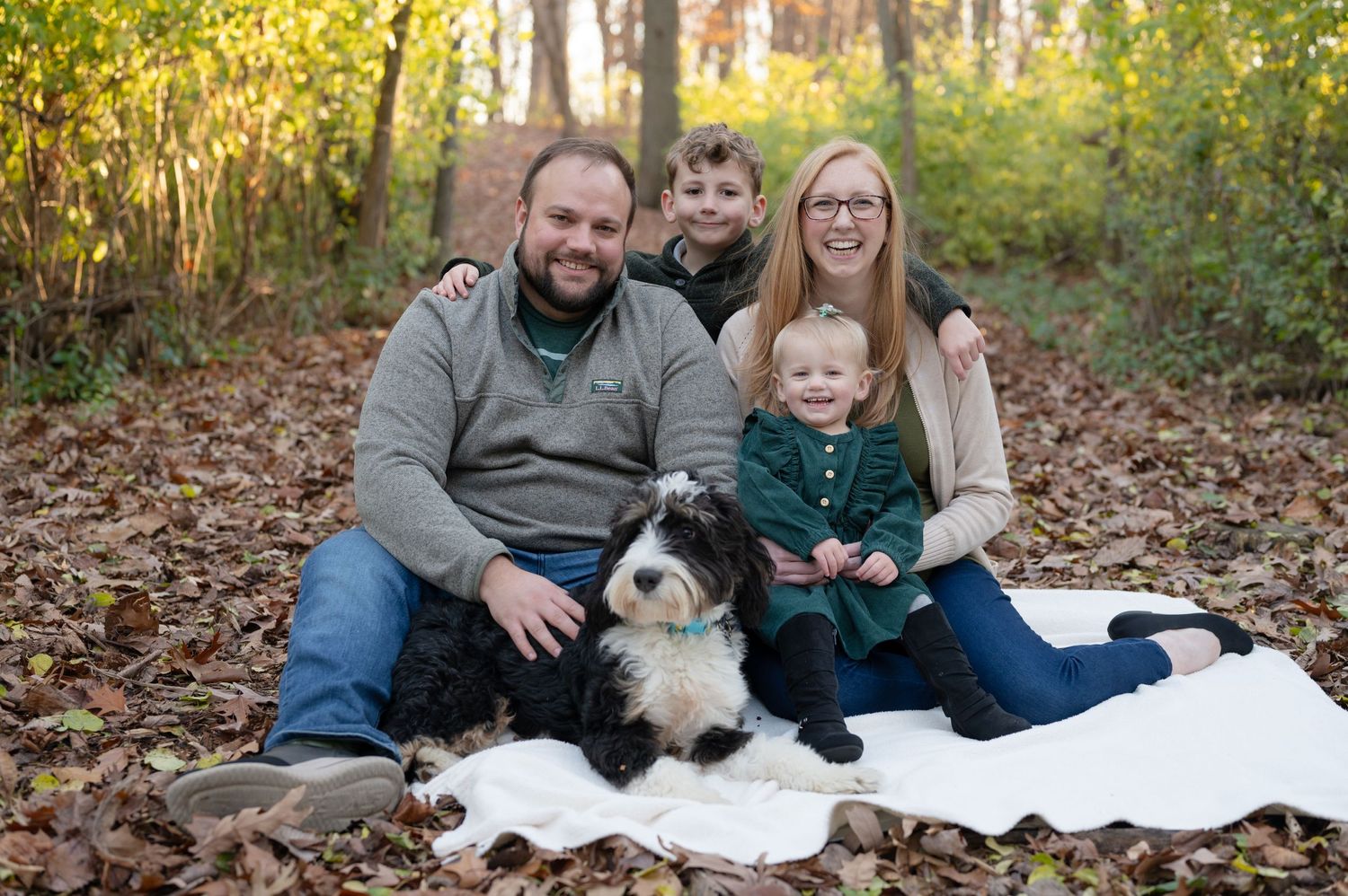 Family sitting on white blanket with their dog in autumn forest setting.