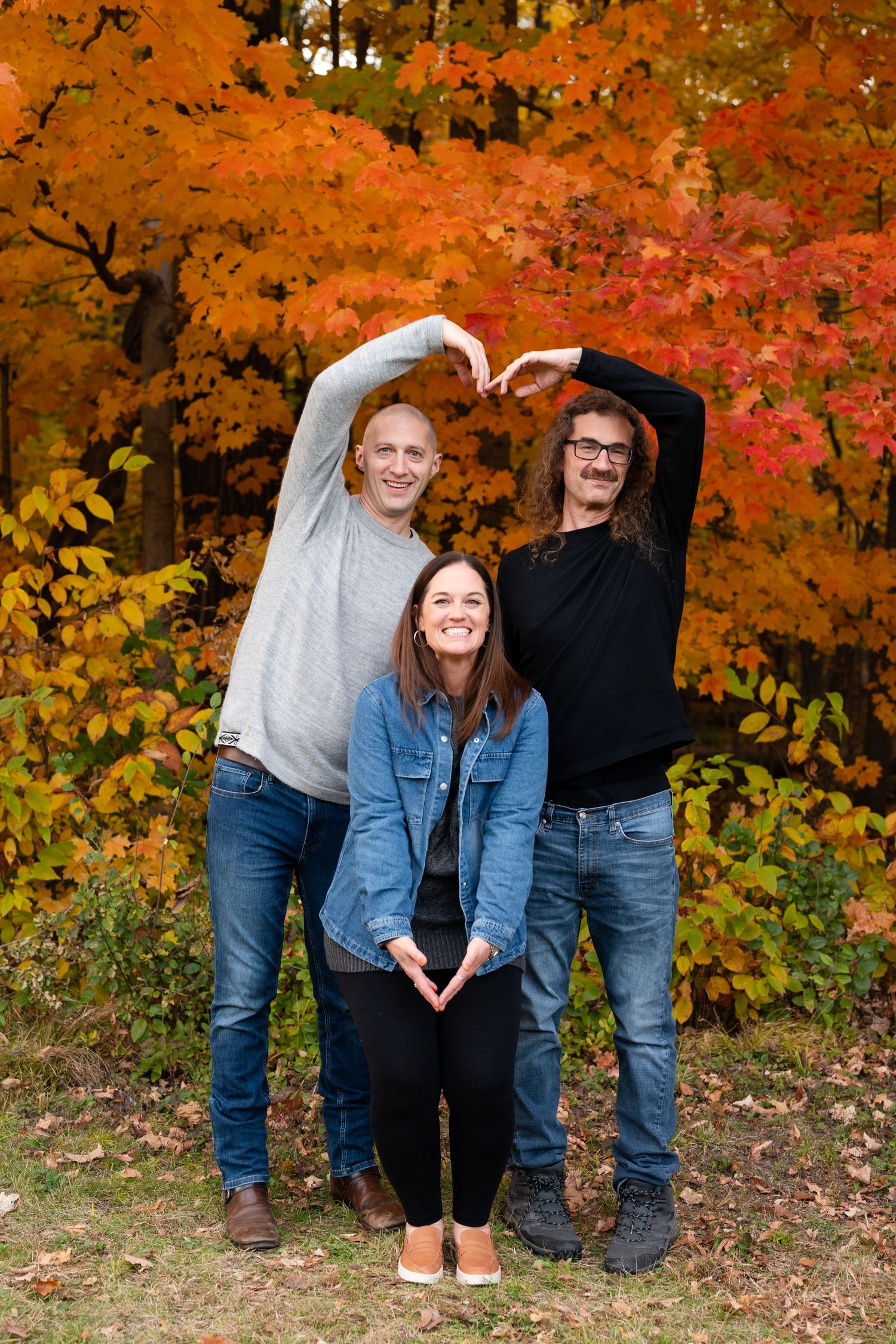 Three people making heart shapes with their arms against vibrant fall foliage.
