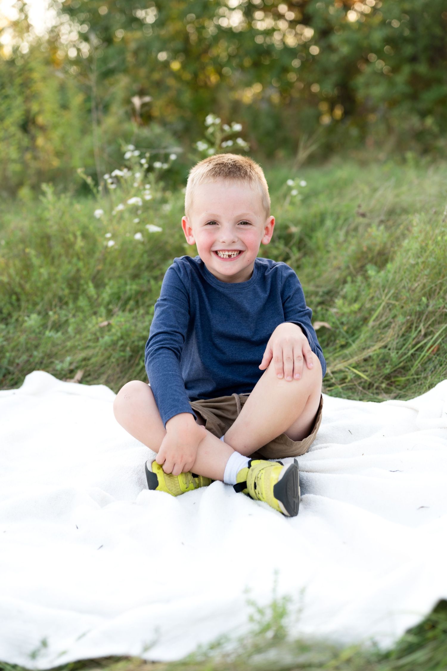 A child in a navy blue sweater sits on a white blanket outdoors surrounded by grass and natural scenery.