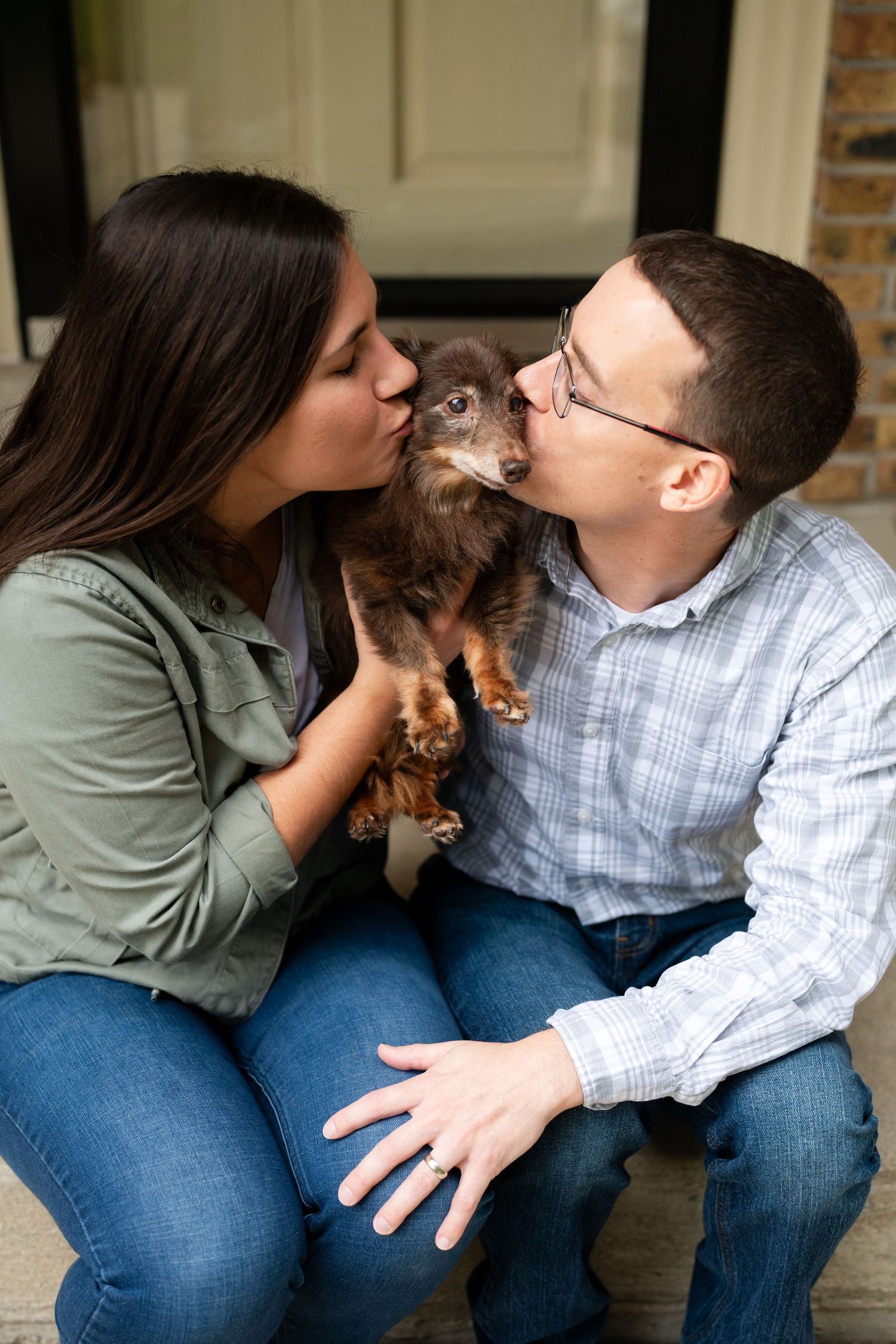 Couple kissing their small brown dachshund while sitting on front porch steps.