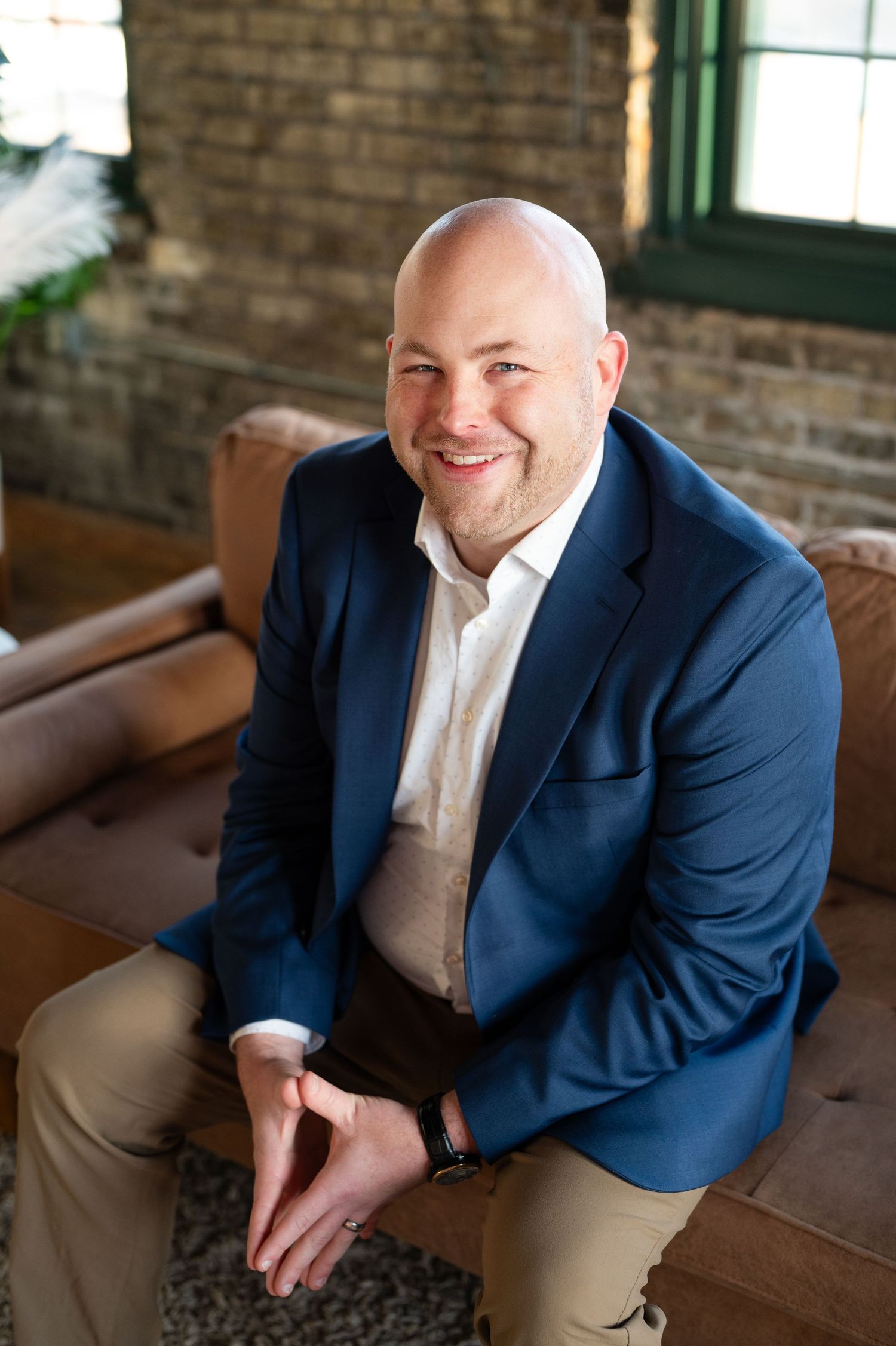 Professional in navy blazer sits relaxed on brown leather couch in rustic brick interior.