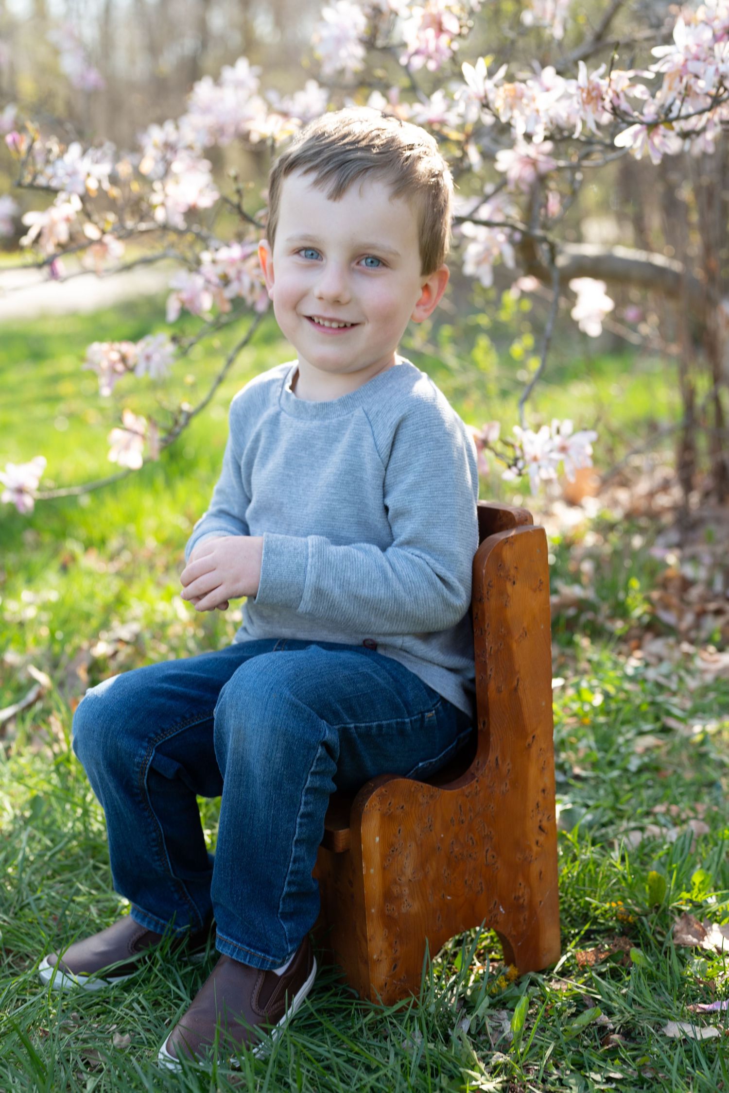 Young child sitting on wooden chair outdoors with cherry blossoms in background.