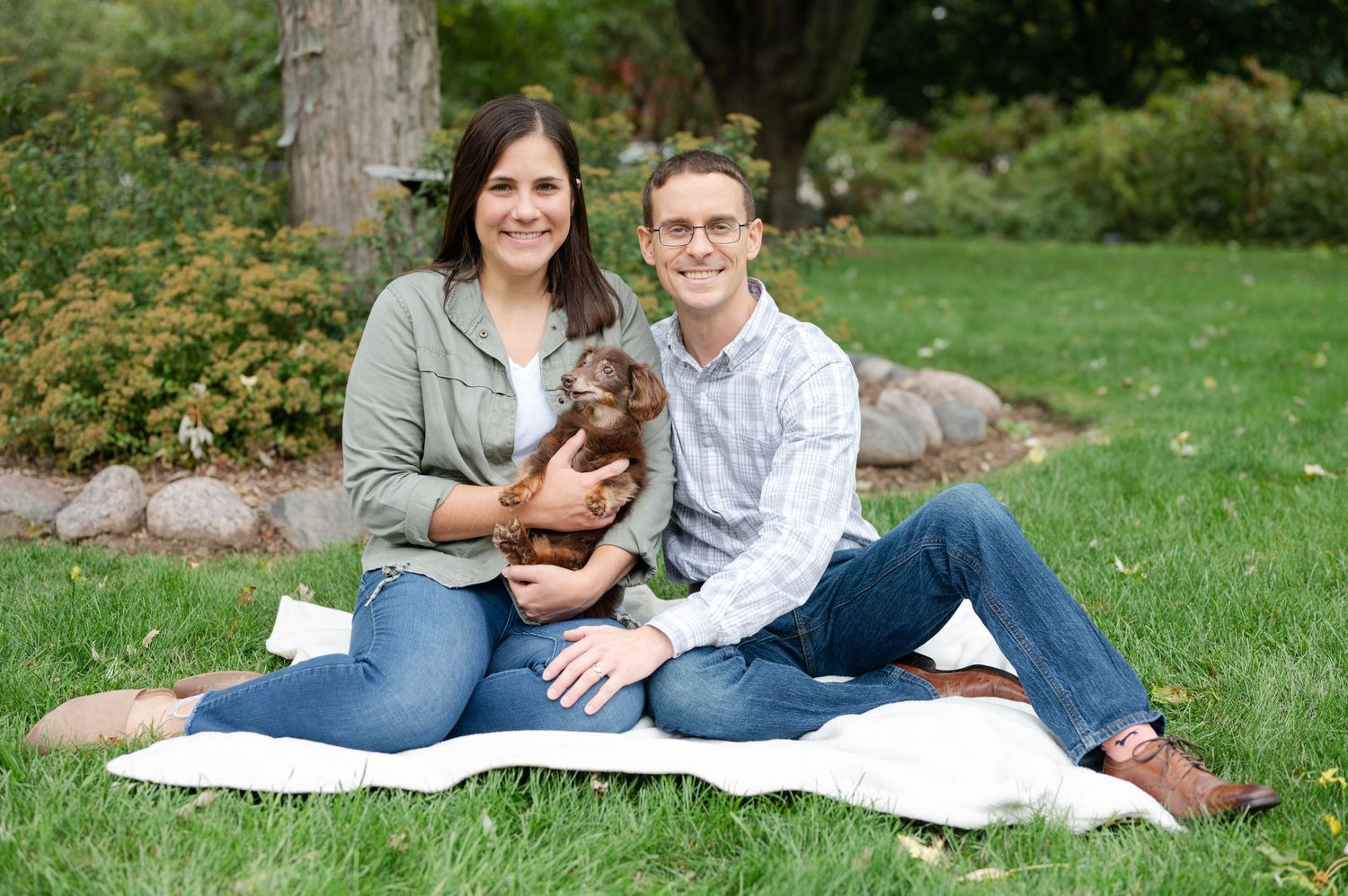 Couple sits on blanket outdoors with their small brown dachshund puppy.
