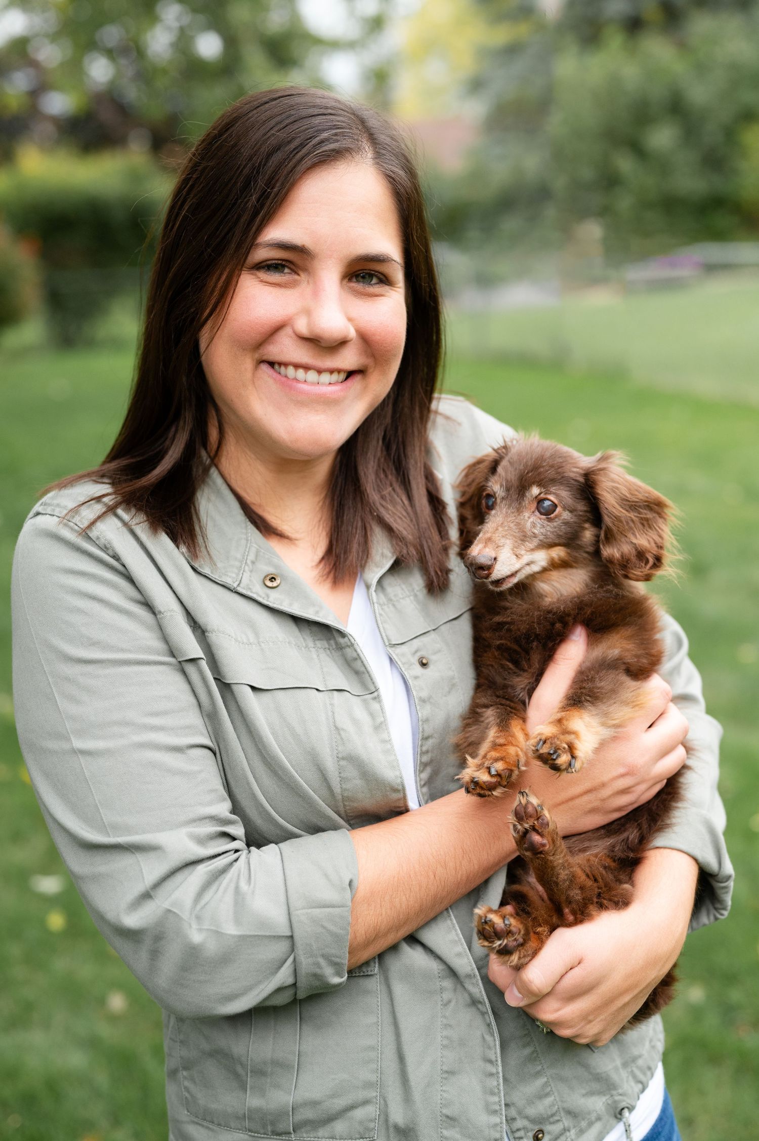 Woman in gray jacket holding small brown dachshund dog outdoors in garden setting.