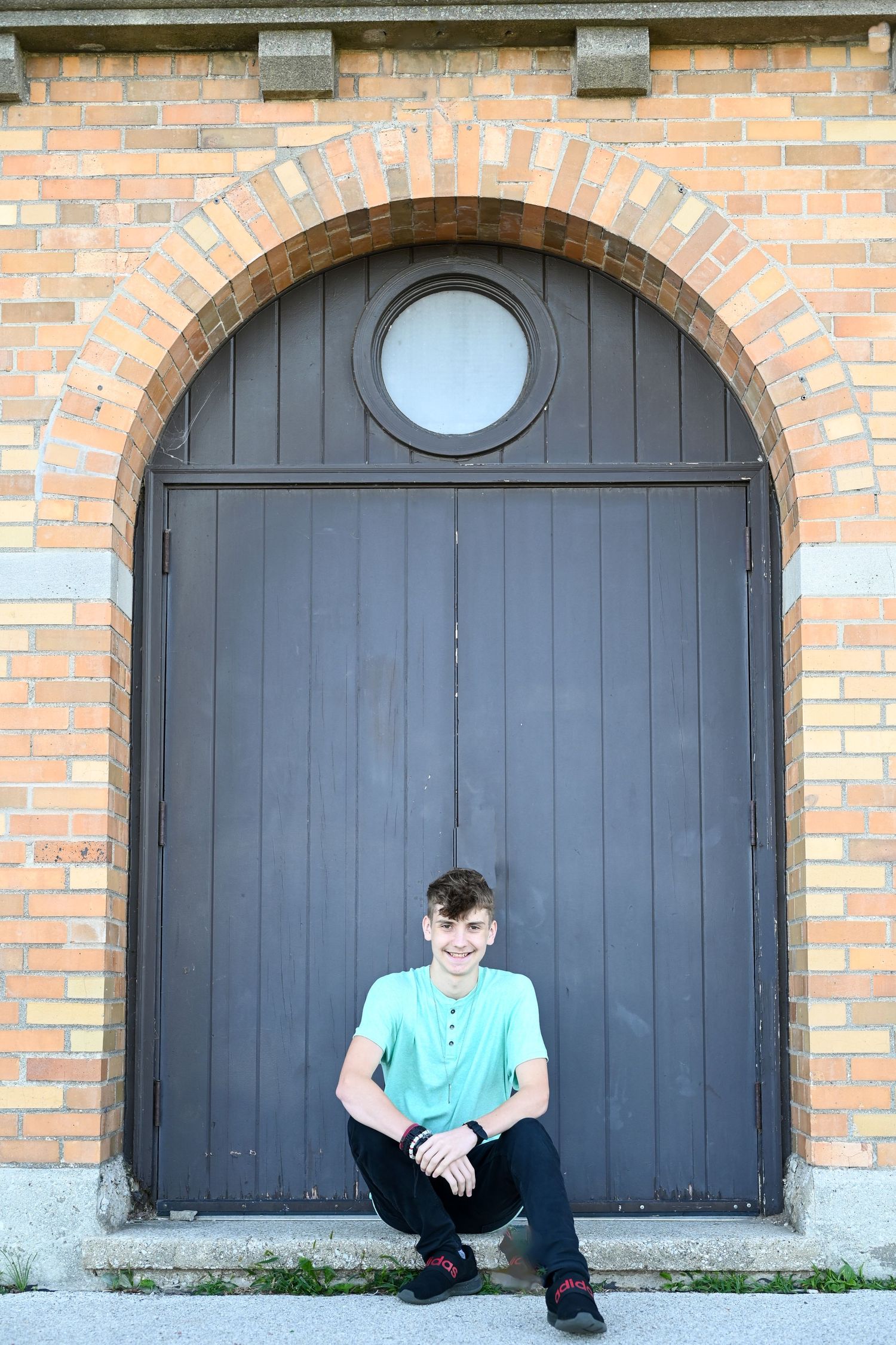 Person sits against a large arched wooden door with circular window in brick building.