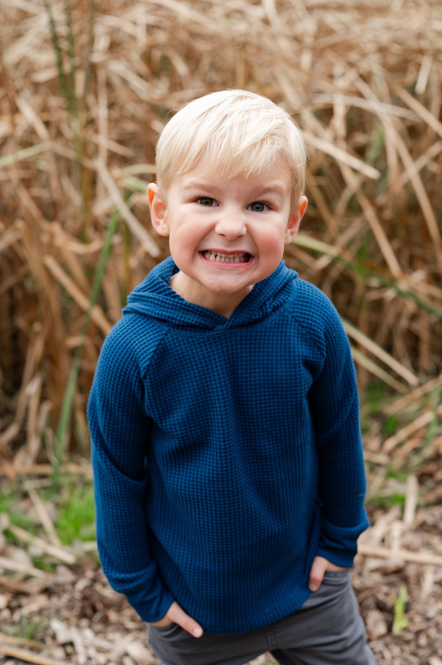 Young child in blue hoodie smiling brightly against backdrop of dried autumn grass.
