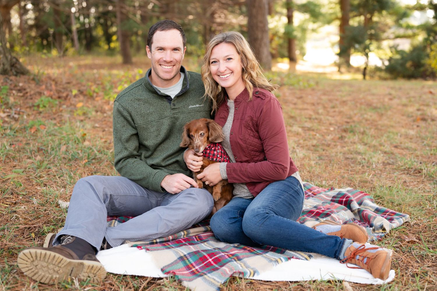 Couple sits with small brown dog on plaid blanket in sunny forest clearing.