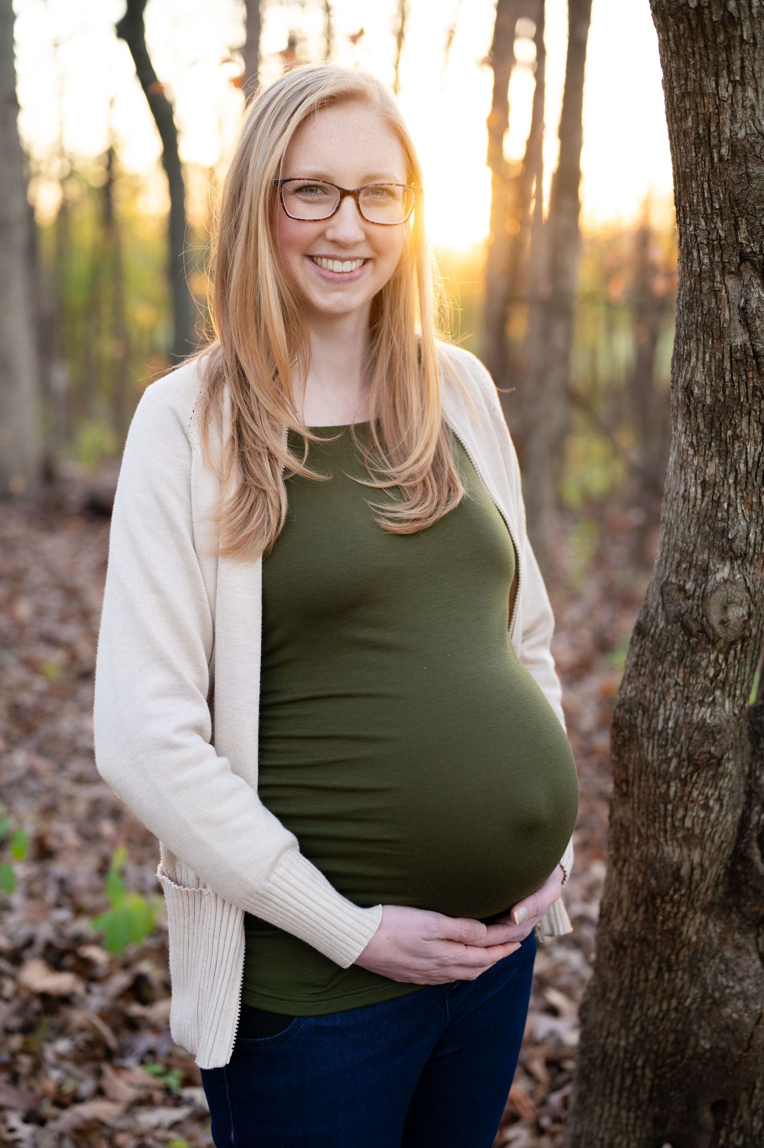 Sunlit maternity portrait in a forest setting wearing a green top and cream cardigan.