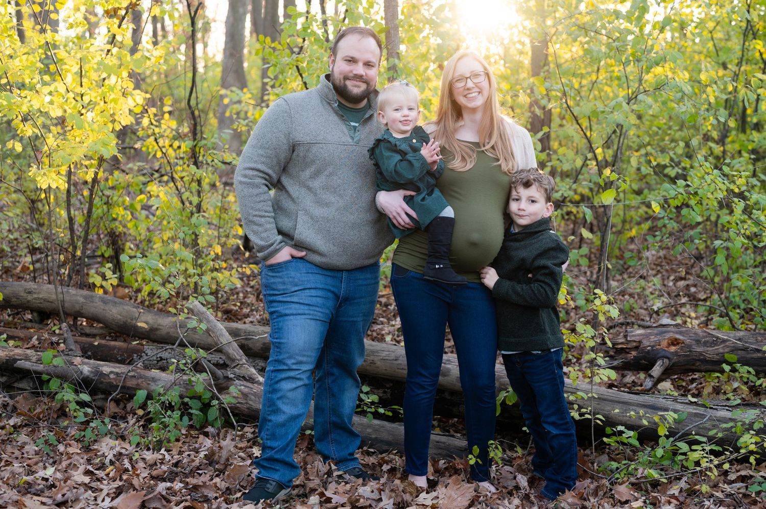Family maternity portrait in a sunlit forest with autumn leaves and natural scenery.