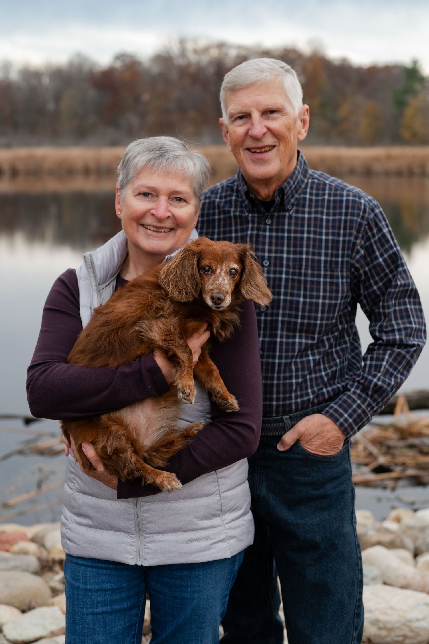 Couple with their brown dachshund dog posing by a lake on an autumn day.