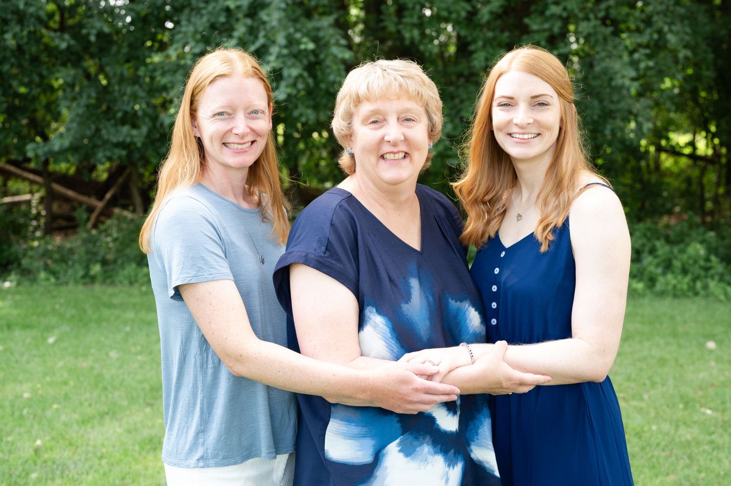 Three people in blue and navy outfits posing together on a sunny day with trees in background.