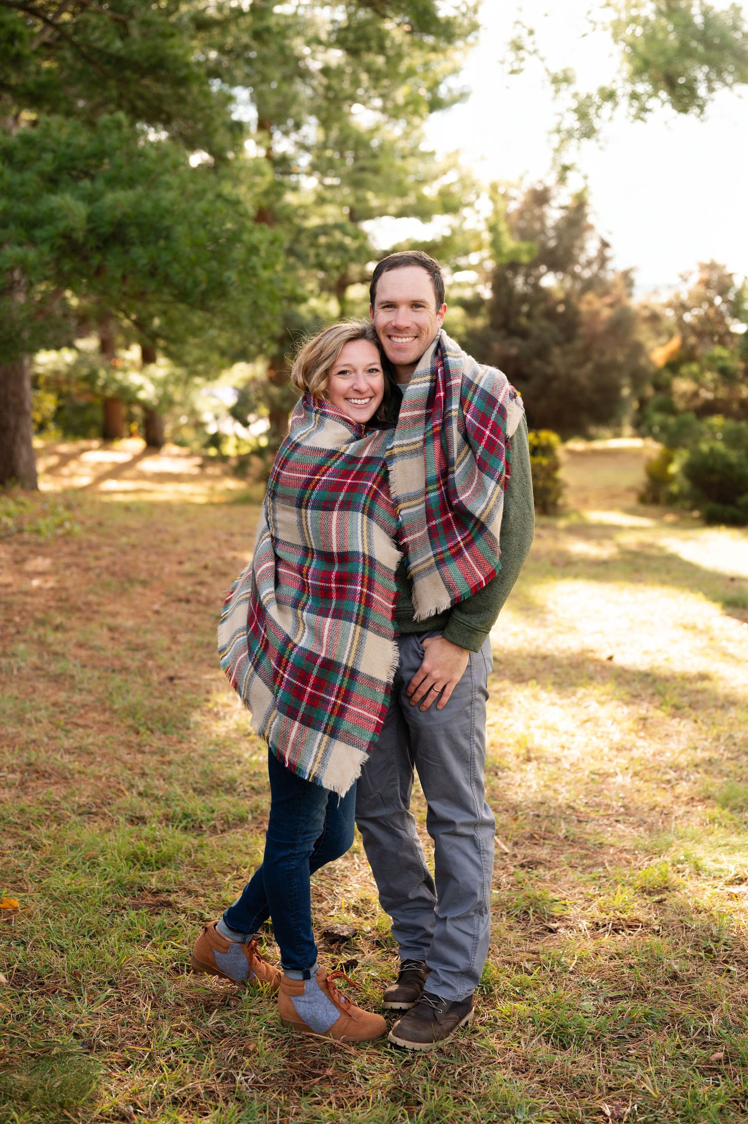 Couple wrapped in plaid blanket embraces while standing in pine forest.
