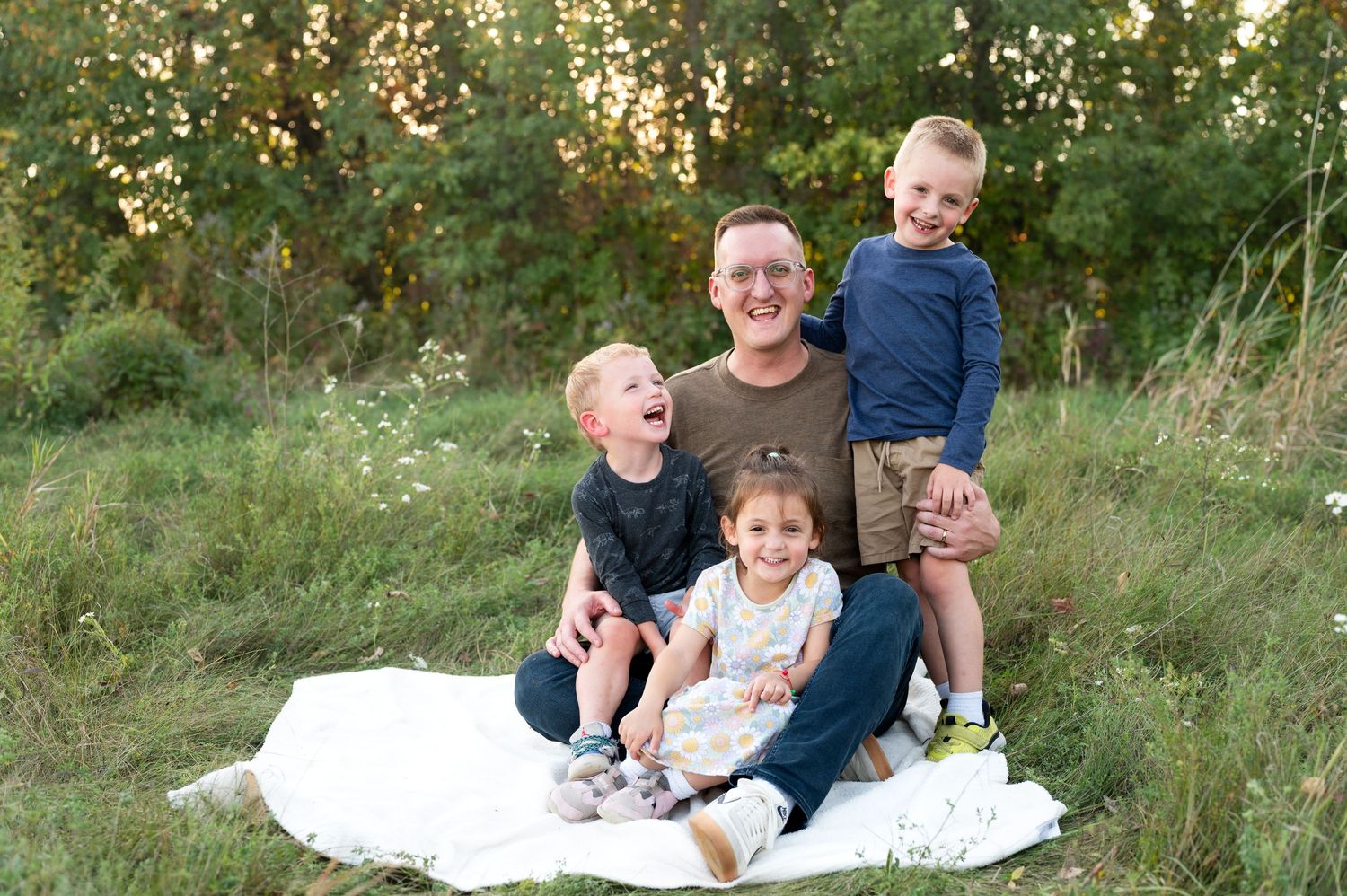 A family laughs together while sitting on a white blanket in a grassy outdoor setting during golden hour.
