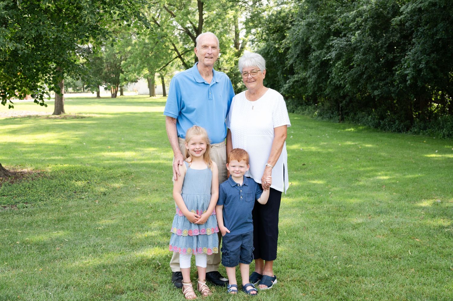 A small family stands together on grass with leafy trees behind them on a bright summer day.