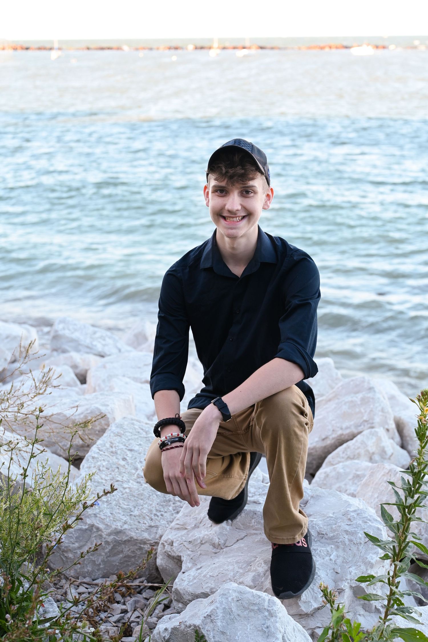 Person in black jacket and baseball cap poses on rocky shoreline with blue water in background.