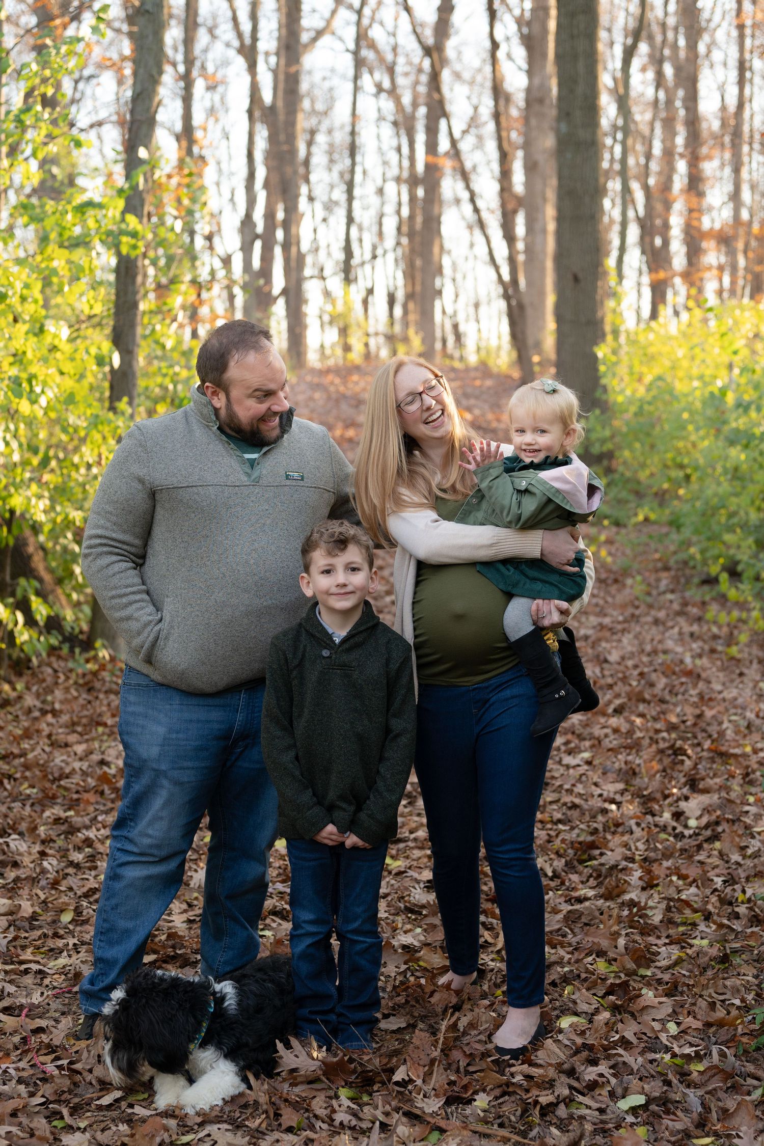 Family of four with their black and white dog posing on fallen leaves in an autumn forest.