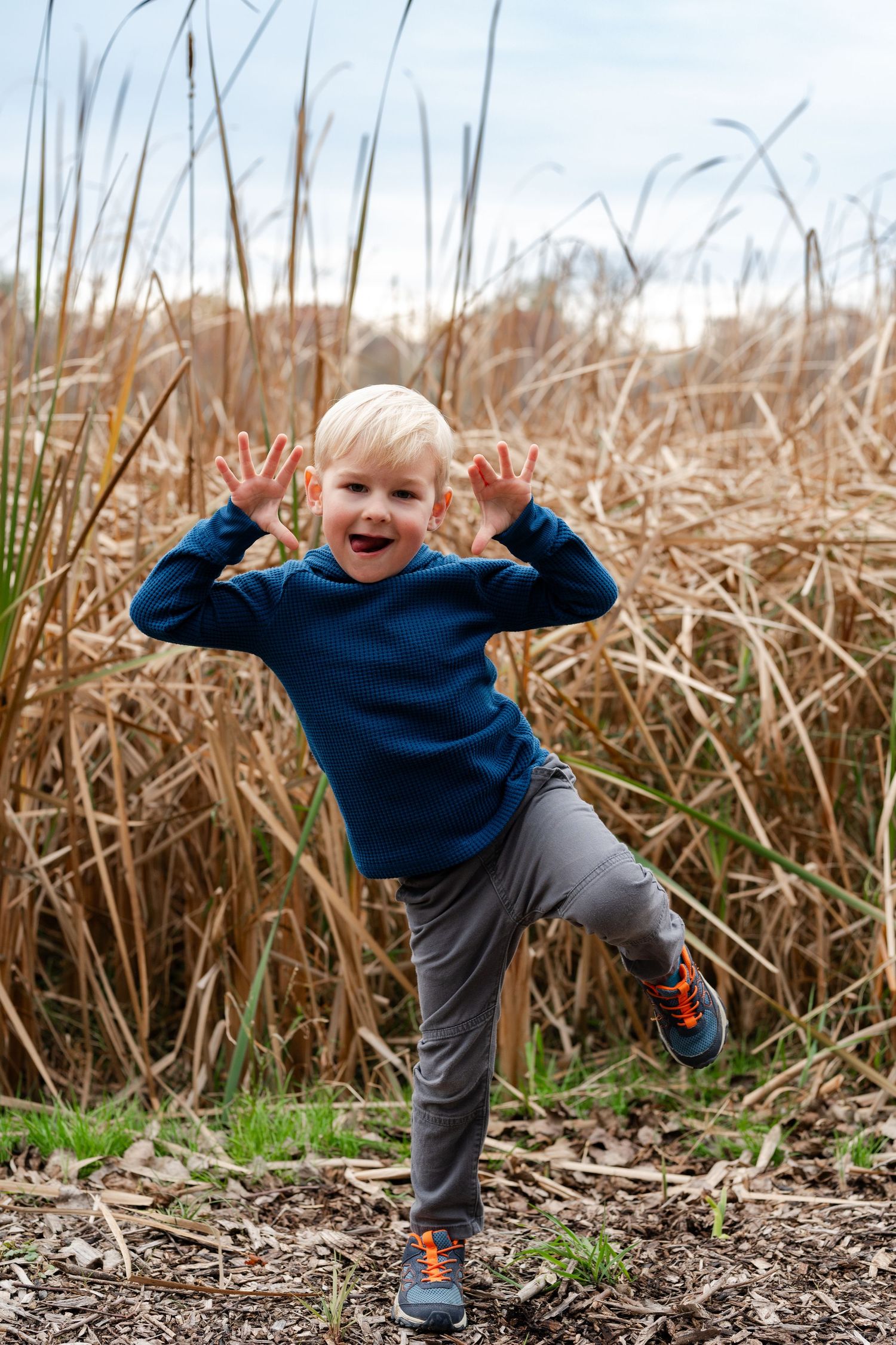 Playful child in blue sweater jumping and making silly faces among tall dried grasses.