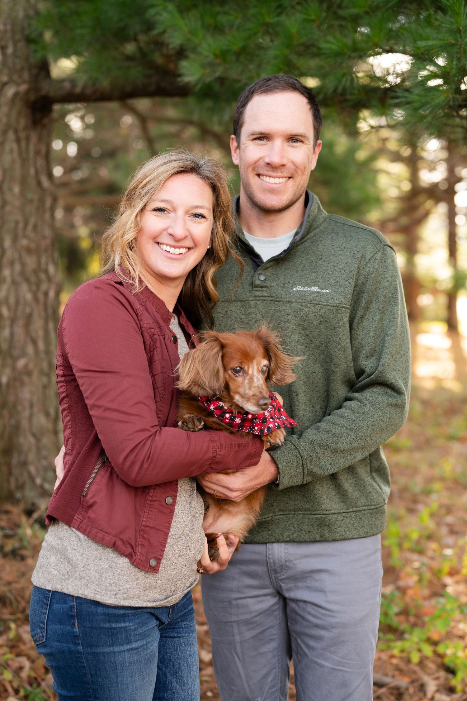 Couple holds small brown dog wearing festive bandana while standing in wooded area.