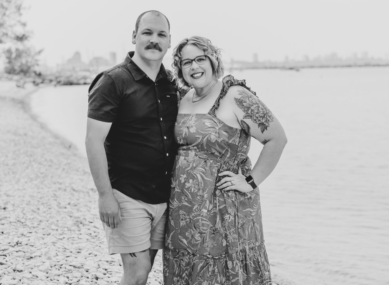 Black and white portrait of a couple by the waterfront in casual attire.