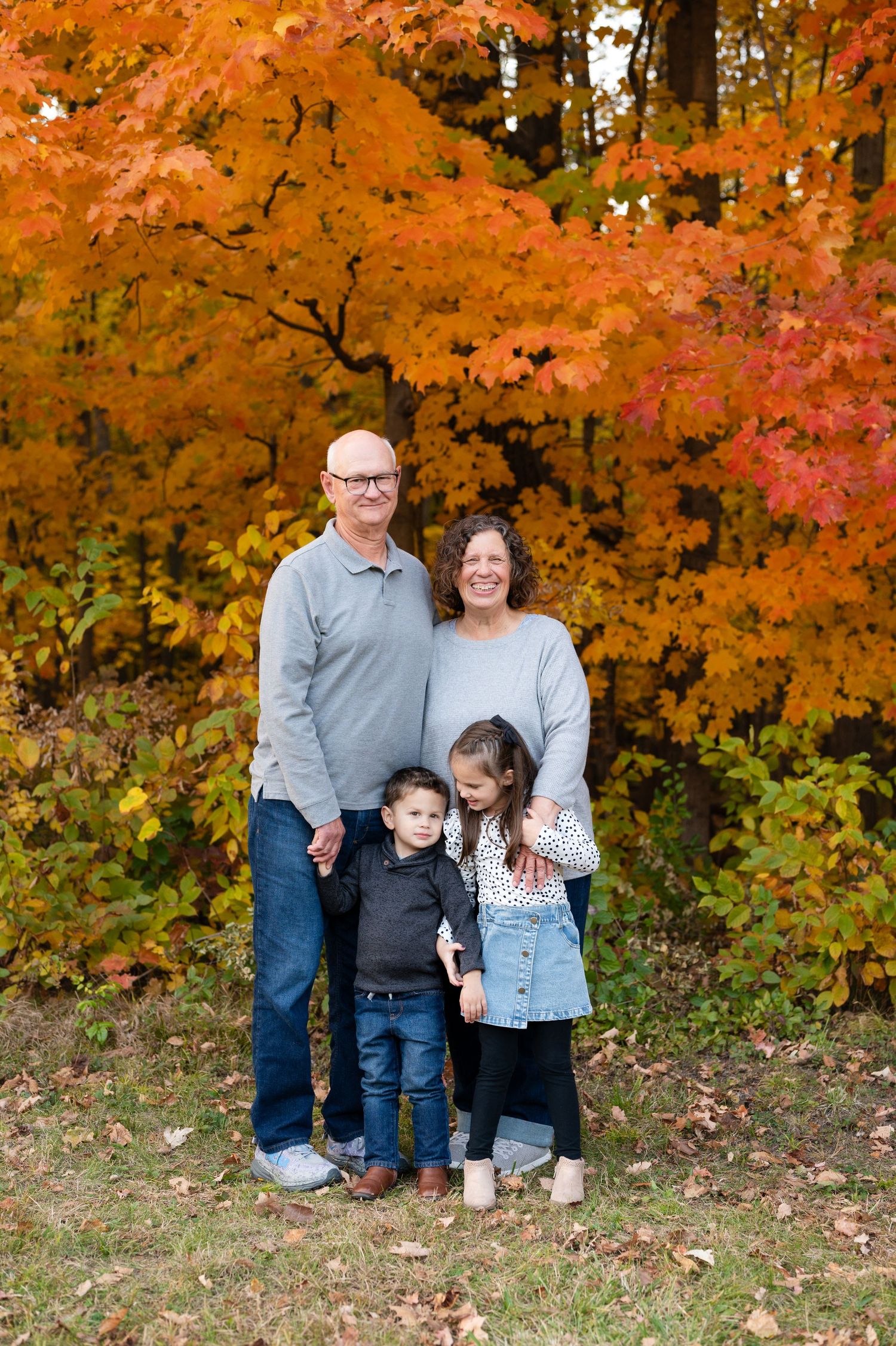 Family of four standing together for autumn portrait with bright orange maple trees in background.