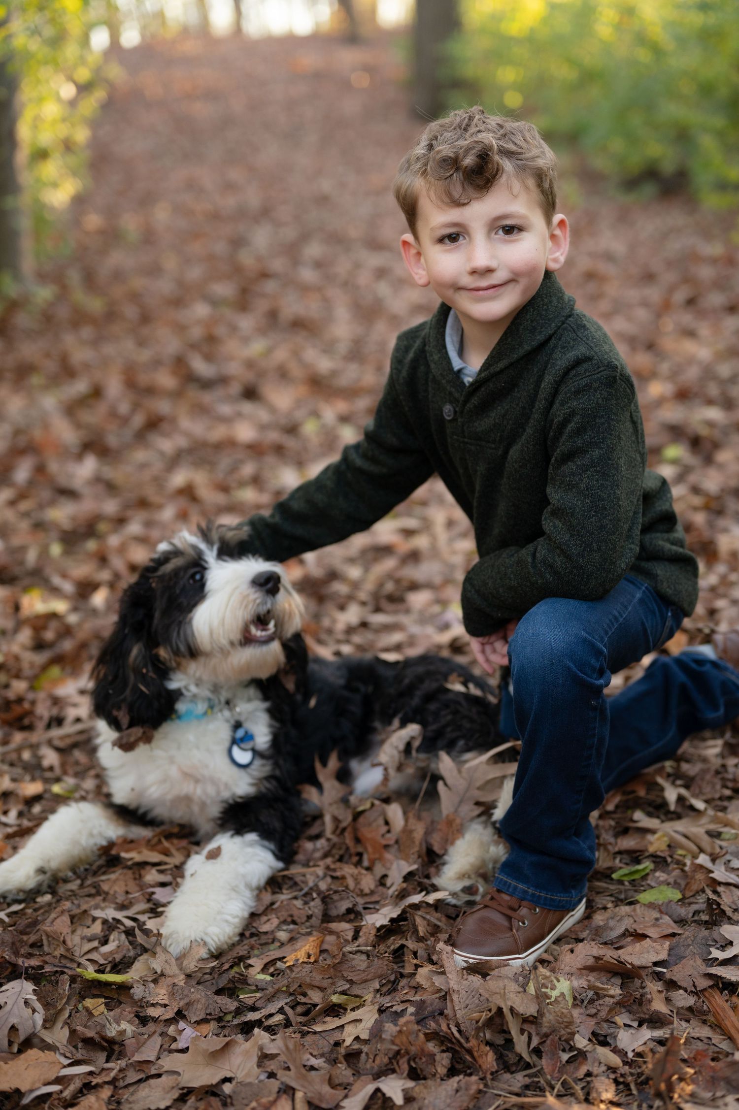 Child kneeling next to black and white dog on fallen autumn leaves.