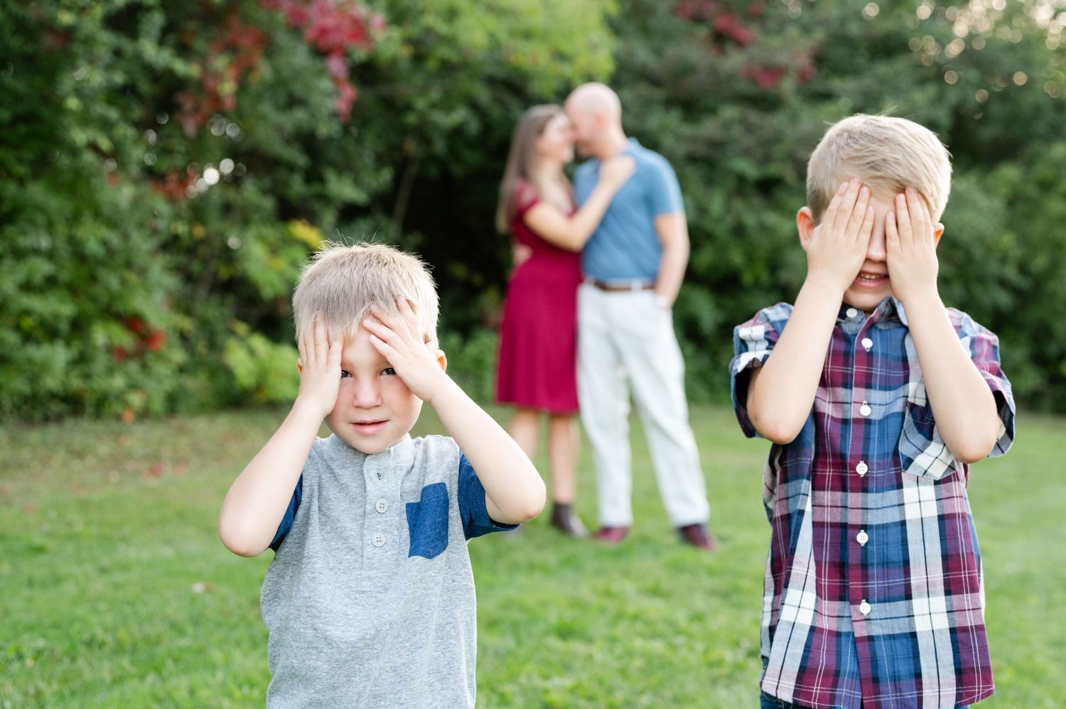 Two children covering eyes while playing outdoors with adults visible in background.