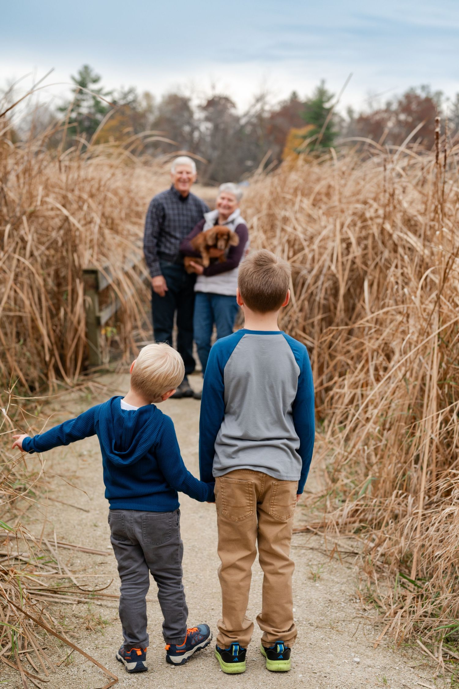 Two children standing on path watching adults with dog in distance among tall grasses.
